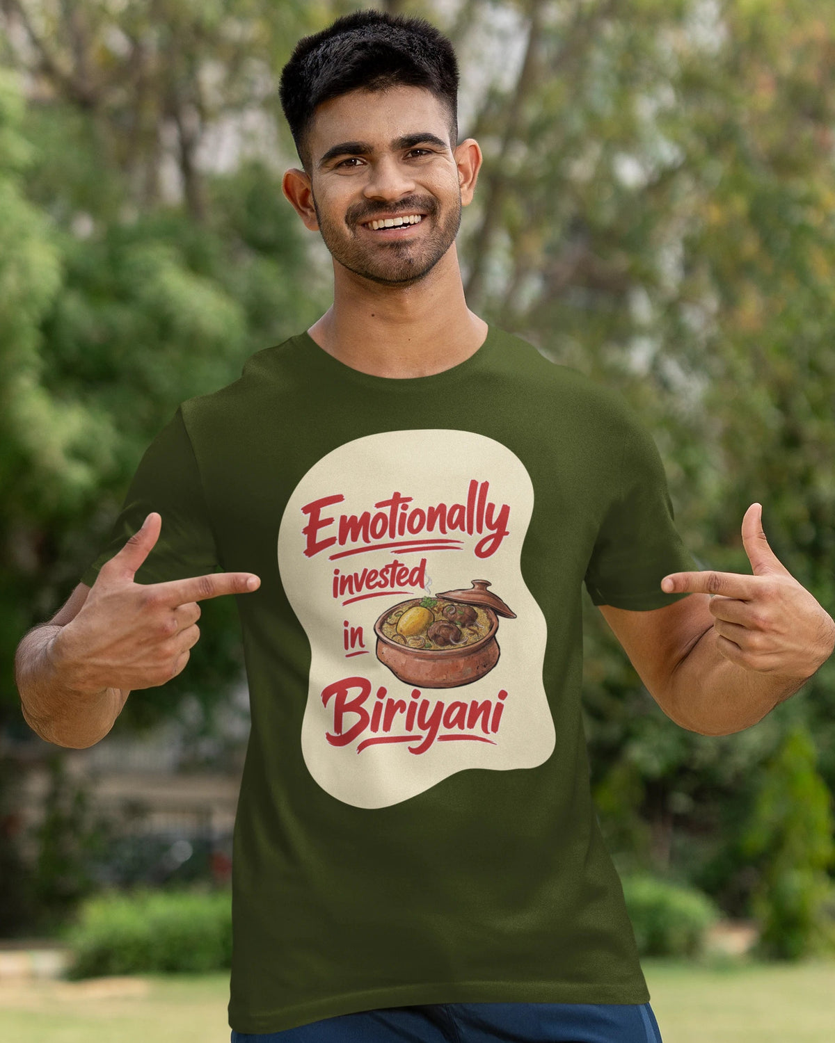 A man wearing a dark olive green t-shirt with a graphic print that reads 'Emotionally invested in Biriyani' in red and cream, posing outdoors with a cricket bat against a blurred green background.