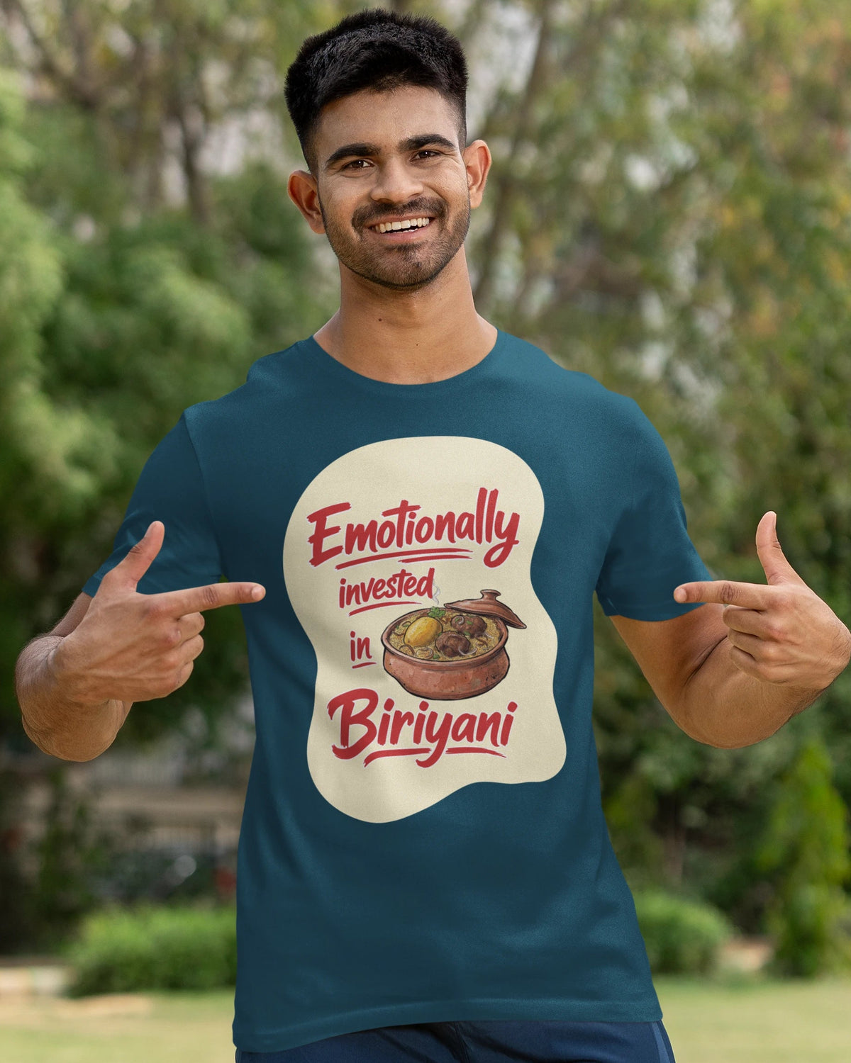 A man wearing a dark teal graphic t-shirt with a 'Emotionally invested in Biriyani' print, standing outdoors in a park-like setting with trees and grass in the background.