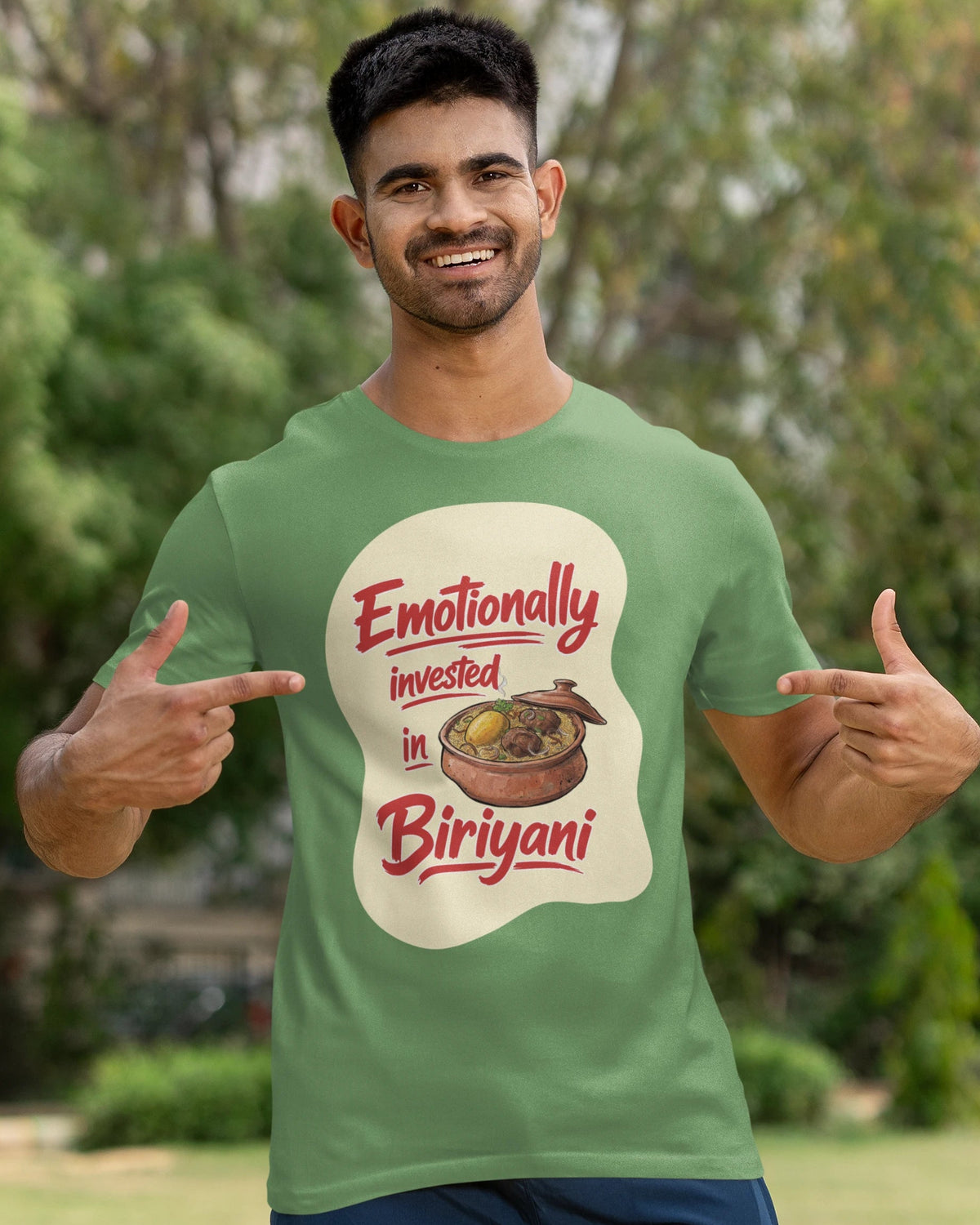 A man wearing a green graphic t-shirt with a 'Emotionally invested in Biriyani' print, posing outdoors with a cricket bat on a grassy field.