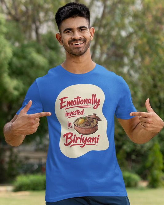 A man wearing a bright blue graphic t-shirt with a white and red circular design that reads 'Emotionally invested in Biriyani' with an illustration of a biriyani dish. He is posing outdoors in a park-like setting with trees and grass in the background.