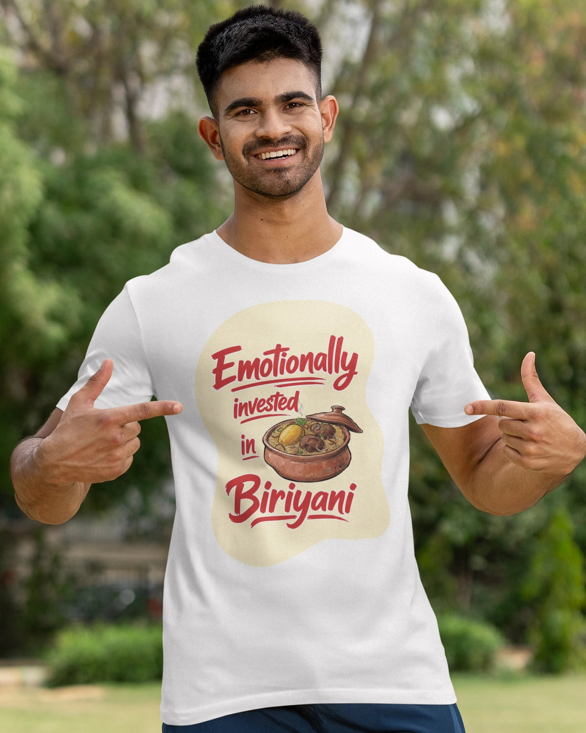 A man wearing a white graphic t-shirt with a 'Emotionally invested in Biriyani' print, standing outdoors in a park-like setting with trees and grass in the background.