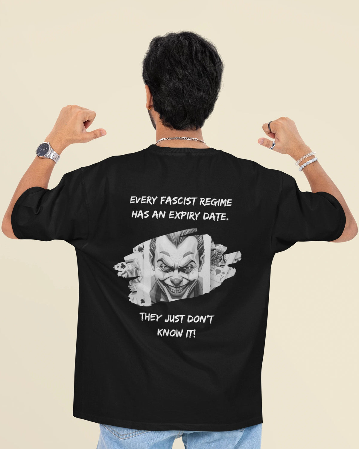 A man shown from behind wearing a black graphic t-shirt with a white printed slogan and clown illustration on the back, paired with light blue jeans, against a solid beige background.