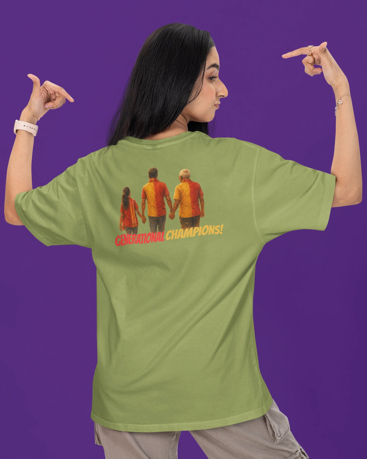 A woman shown from the back wearing an oversized sage green t-shirt with a graphic of three people holding hands and the text 'CHAMPIONS CHAMPIONS!' in orange and red. She is posing with her hands pointing outward against a solid purple background.