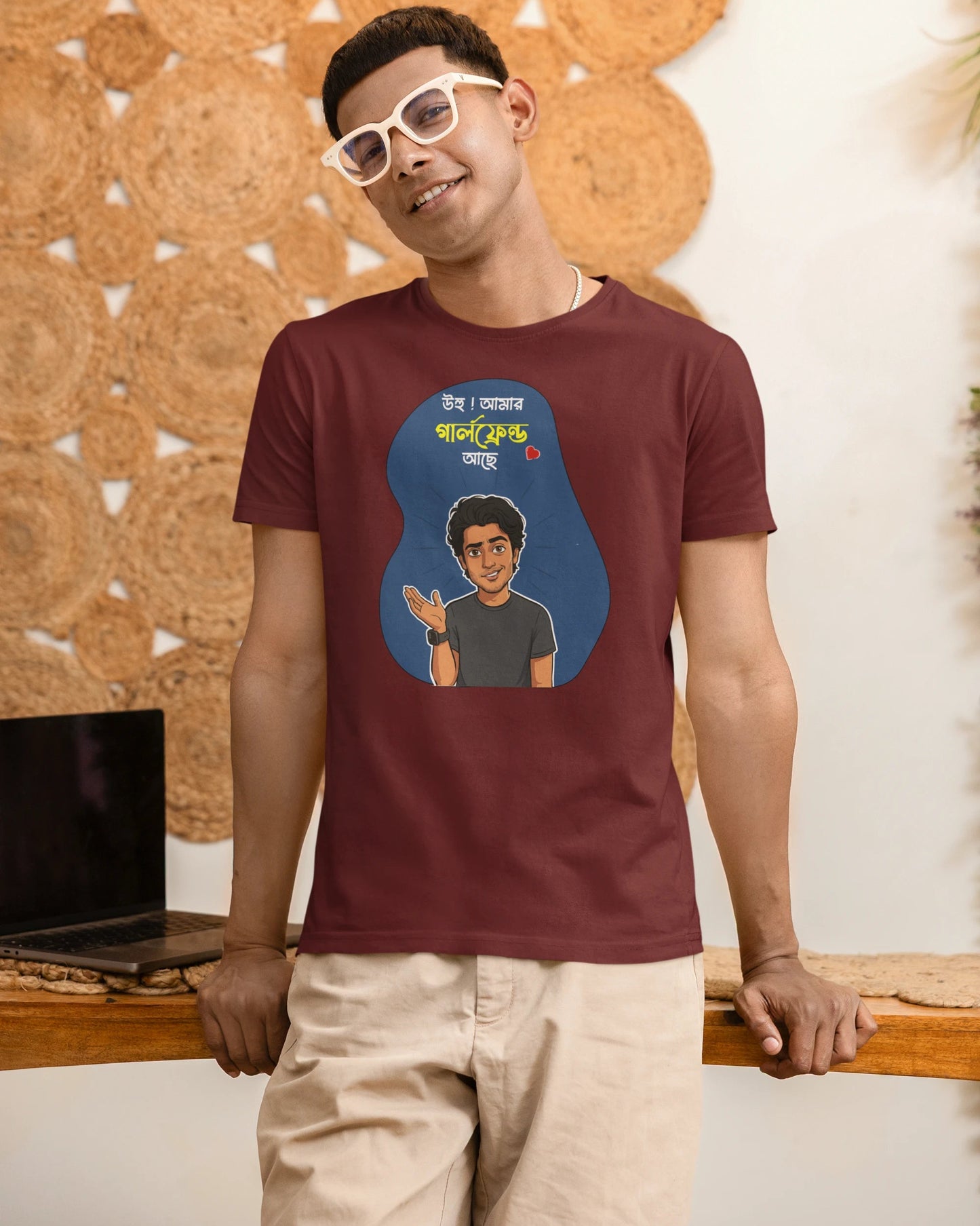 A man wearing a maroon t-shirt with a cartoon graphic and Hindi text, paired with beige pants and white sunglasses, standing in front of a decorative cork wall.