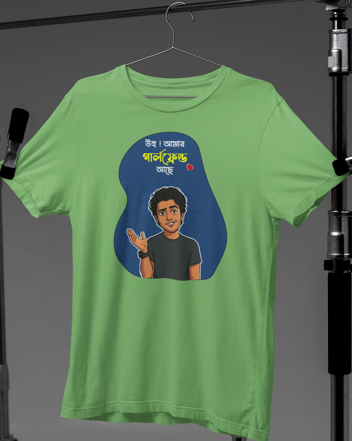 A light green short-sleeve t-shirt hanging on a hanger against a dark gray studio backdrop, featuring a blue graphic with a cartoon portrait and Bengali text on the front.