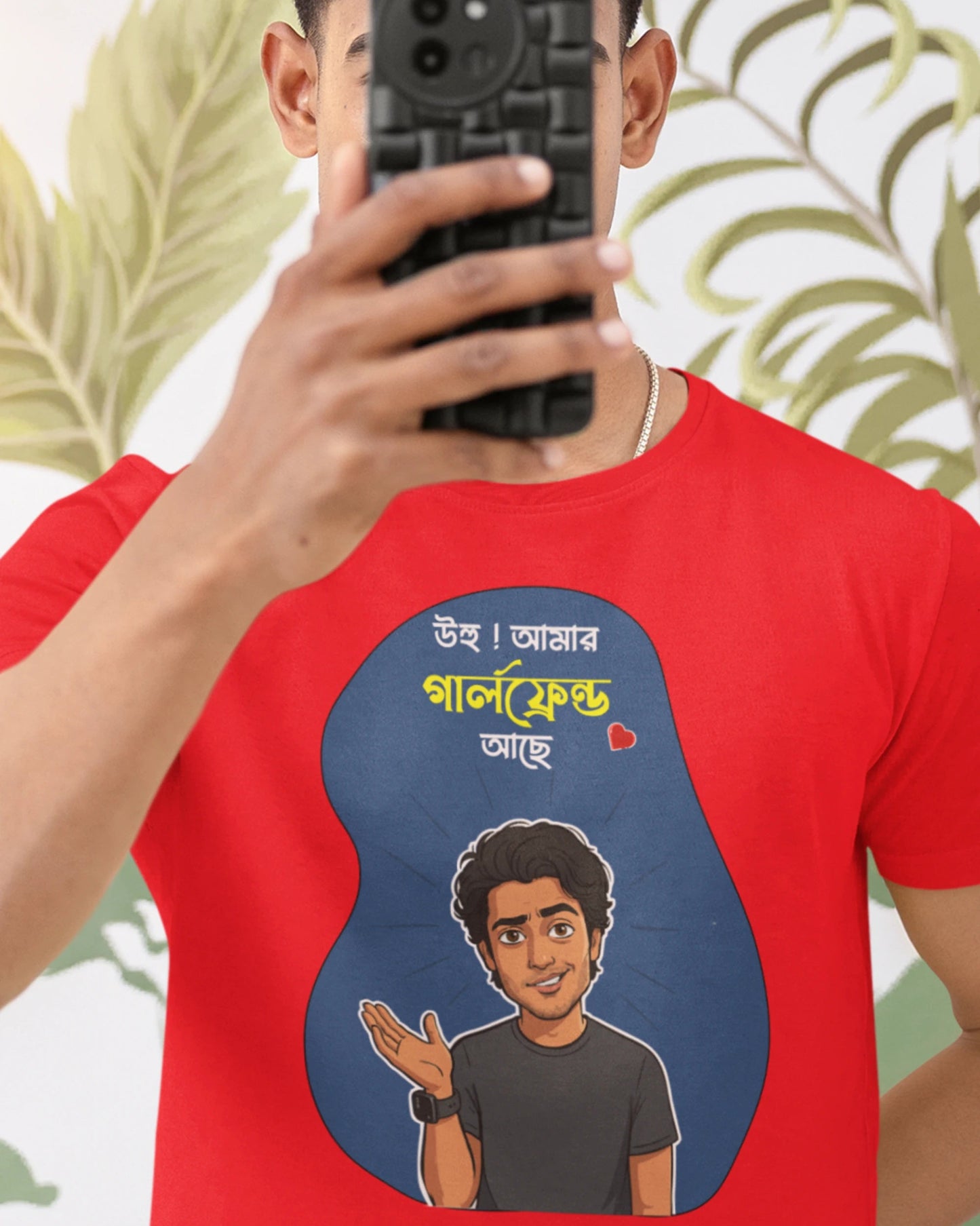A man taking a mirror selfie wearing a bright red t-shirt with a cartoon graphic of a man and Bengali text inside a blue shape. The background is white with green leaf patterns.