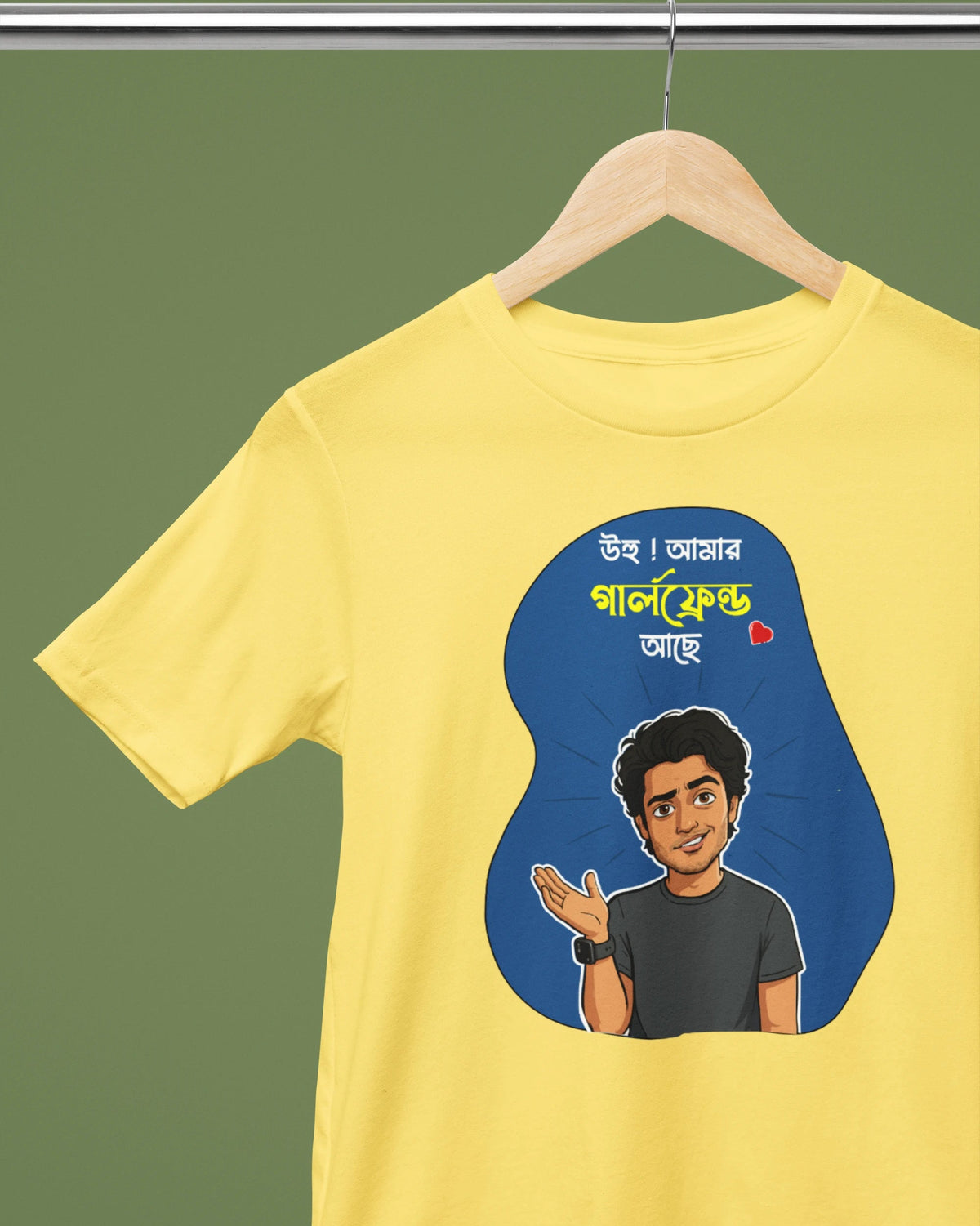 A yellow short-sleeve t-shirt hanging on a wooden hanger against a solid olive green background. The shirt features a graphic print with a cartoon illustration of a man and Bengali text inside a blue abstract shape.