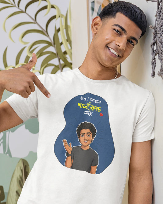 A smiling young man wearing a white graphic t-shirt with a blue abstract shape containing a cartoon illustration and Bengali text. He is pointing at the graphic with his right hand, standing in a room with a light-colored wall and decorative plant leaves.