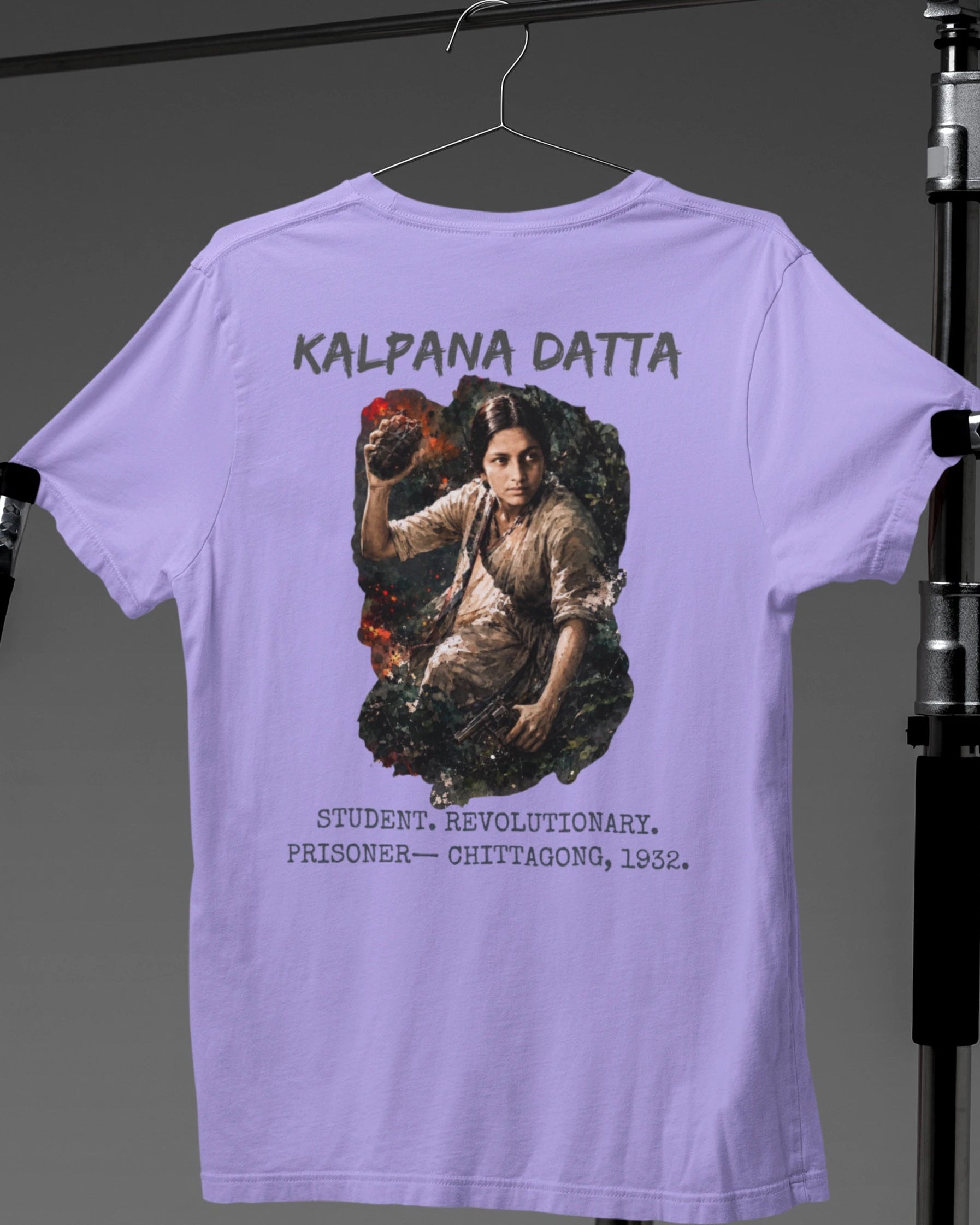 A light purple t-shirt hanging on a hanger, showing the back side with a large graphic print of a woman and the text 'KALPANA DATTA' and 'STUDENT, REVOLUTIONARY, PRISONER - CHITTAGONG, 1932.' The shirt is photographed in a studio setup with lighting equipment visible on both sides.