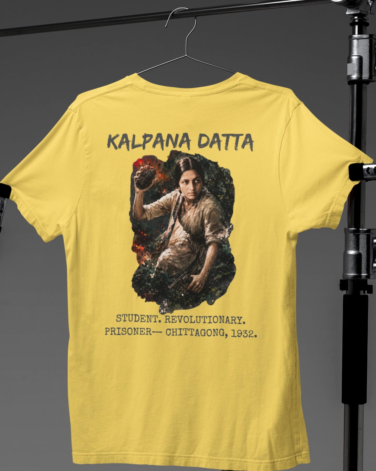 A yellow short-sleeve t-shirt hanging on a hanger against a dark gray studio backdrop, with a large back graphic featuring the text 'KALPANA DATTA' and a portrait illustration. The shirt is displayed on a stand.