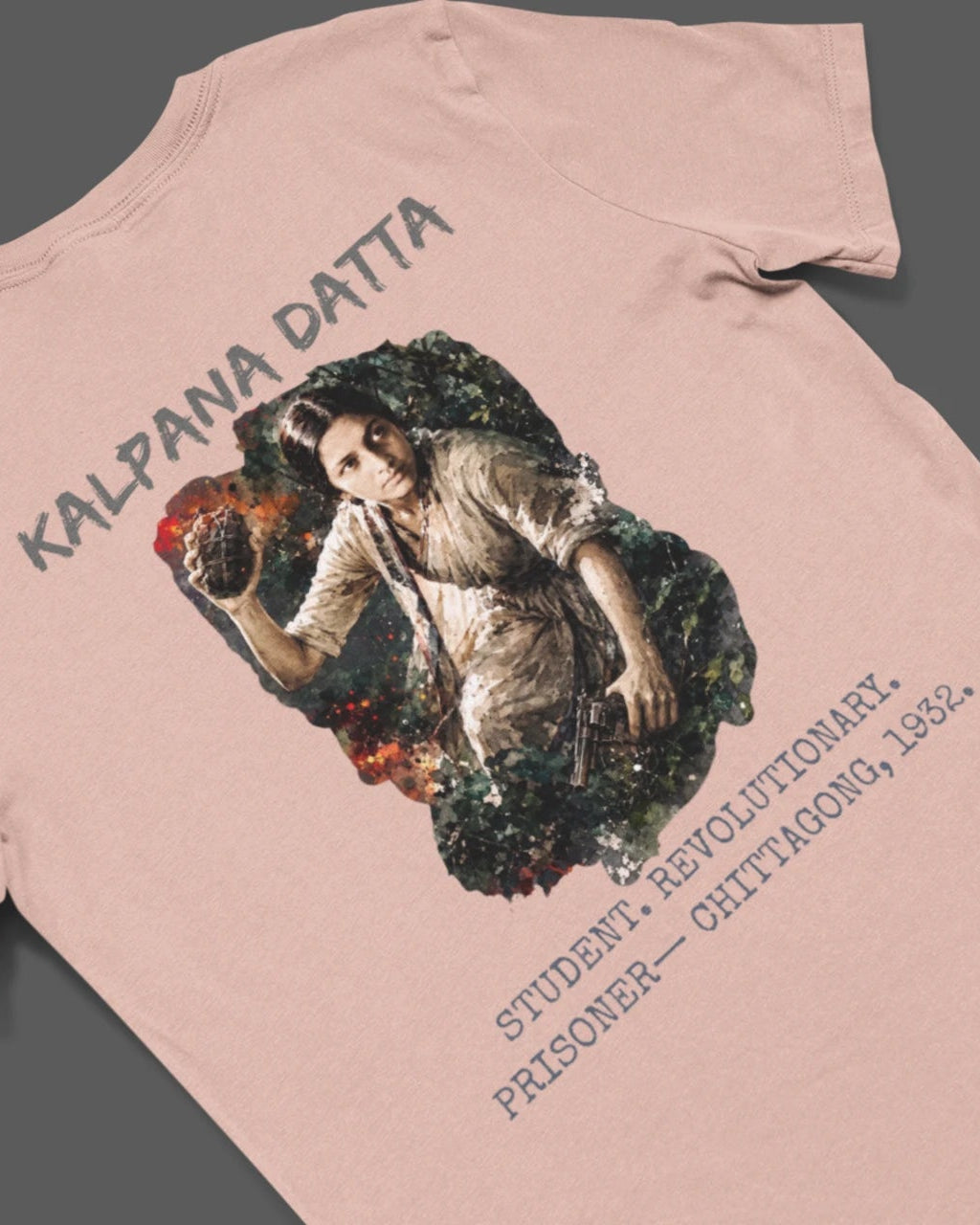 A light pink t-shirt laid flat on a dark gray background, featuring a large graphic print on the back with the text 'KALPANA DATTA' and a classical-style illustration of a woman.