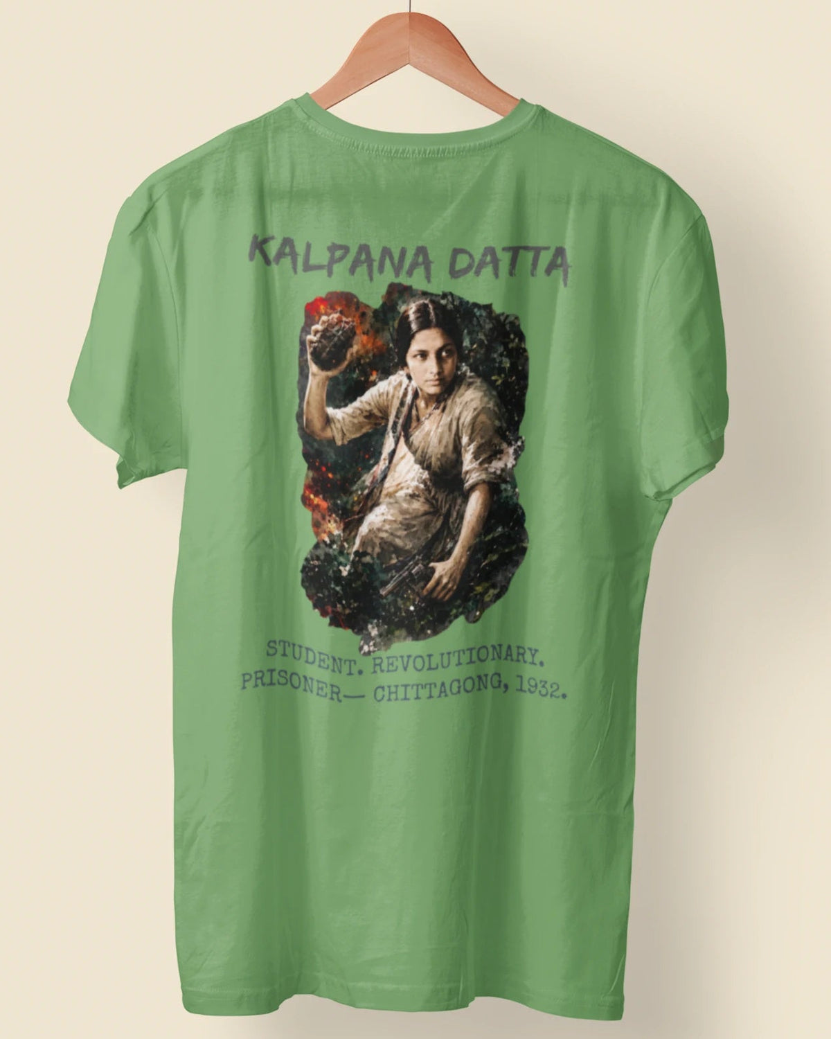 A green short-sleeve t-shirt hanging on a wooden hanger against a light beige background. The back of the shirt features a large graphic print depicting a historical figure with the text 'KALPANA DATTA' above and additional text below.