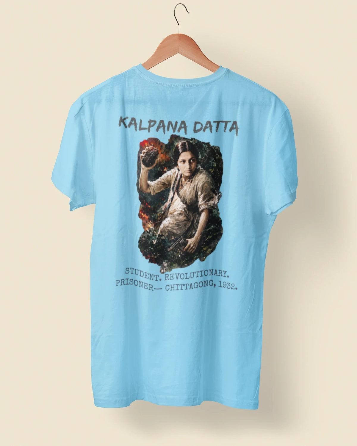 A light blue short-sleeve t-shirt hanging on a wooden hanger against a solid beige background. The back of the shirt features a large graphic print depicting a woman in a white saree surrounded by dark foliage, with the text 'KALPANA DATTA' above and 'STUDENT, REVOLUTIONARY, PREACHER - COTTAYONG 1952.