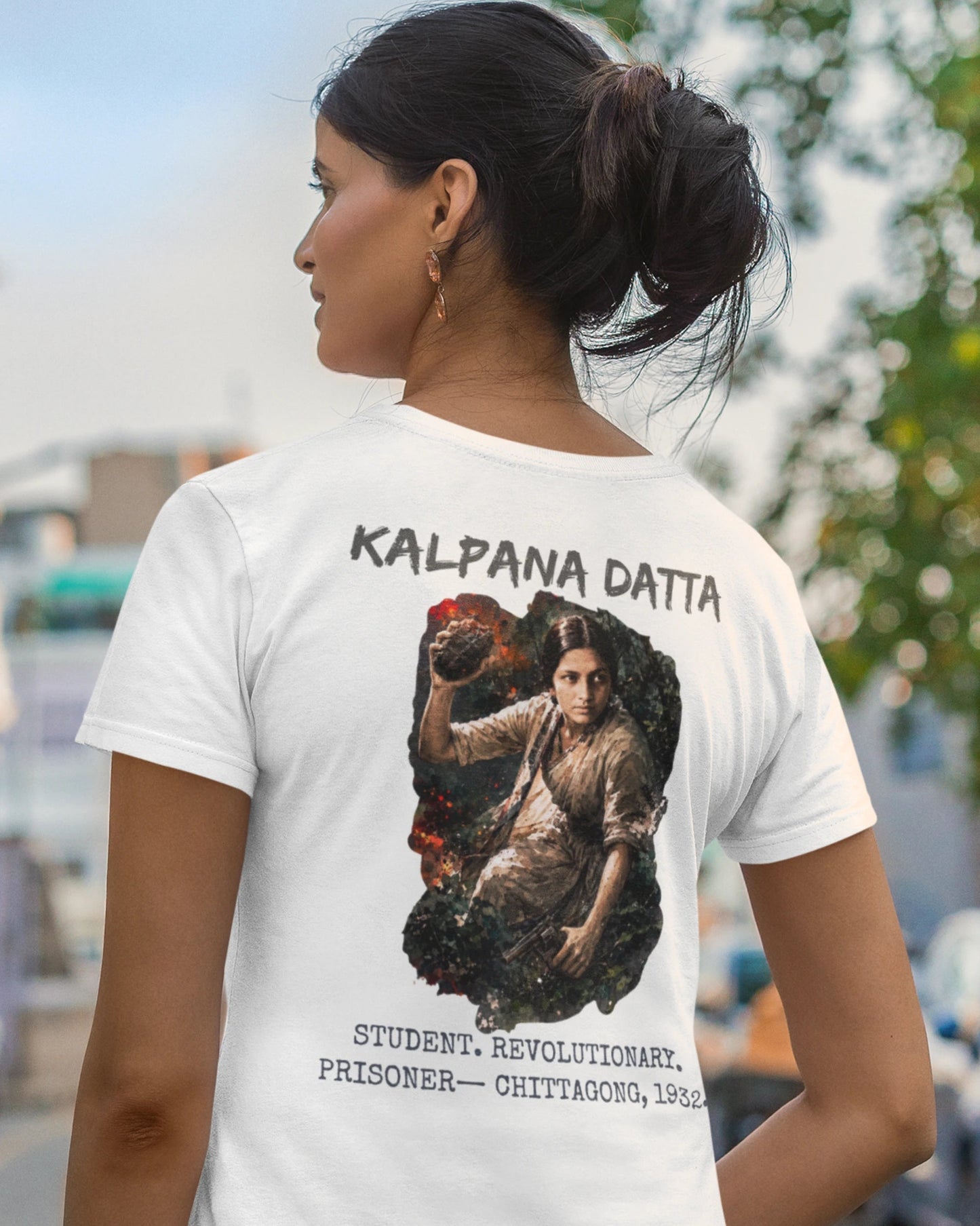 A woman shown from the back wearing a white graphic T-shirt with the text 'KALPANA DATTA' and a large printed portrait and additional text on the back, photographed outdoors on a blurred street background.