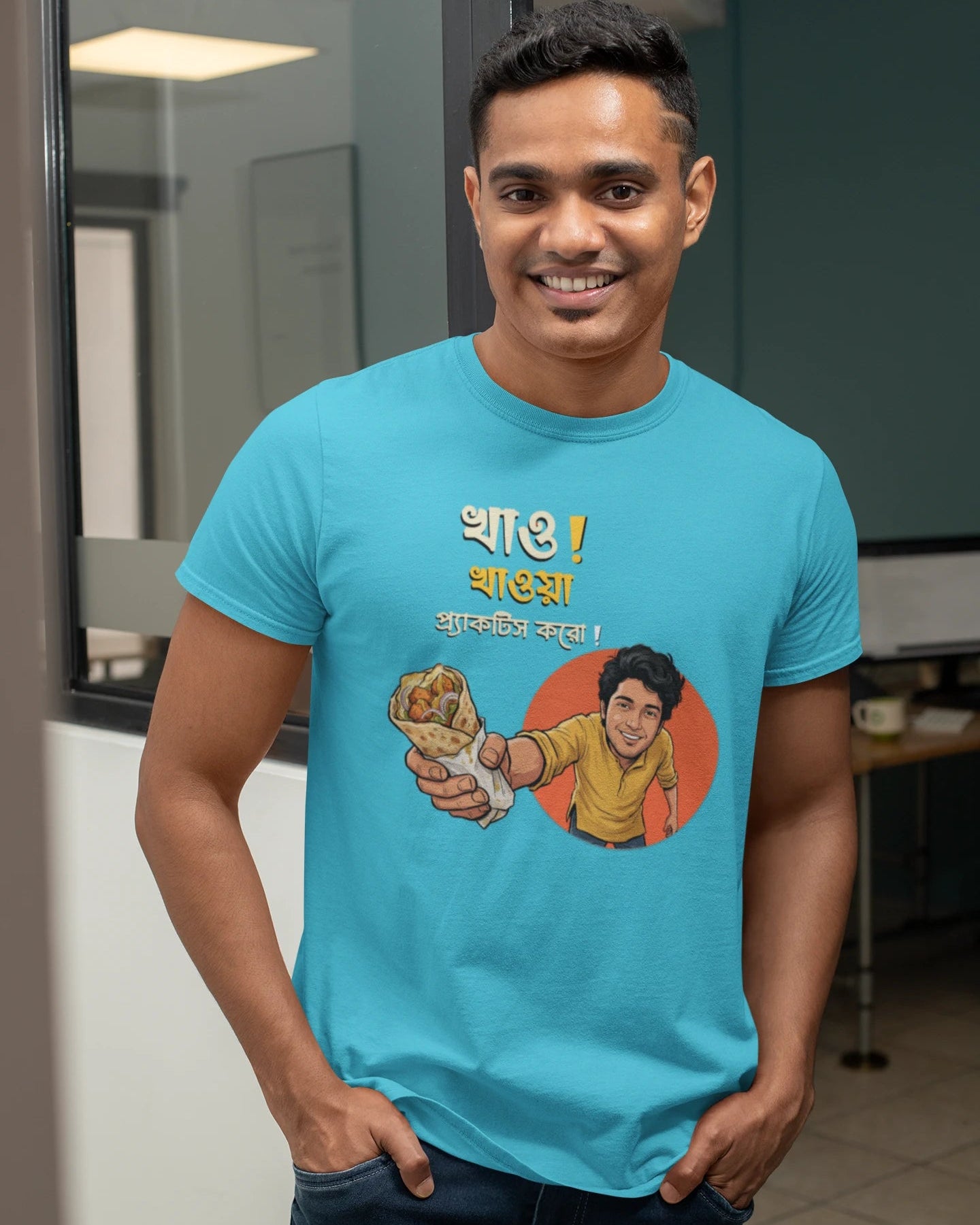 A smiling man wearing a dark "Bong Cazuals" graphic t-shirt featuring Bengali typography and an illustrated character offering a food roll, shot in an indoor office or lifestyle setting.
