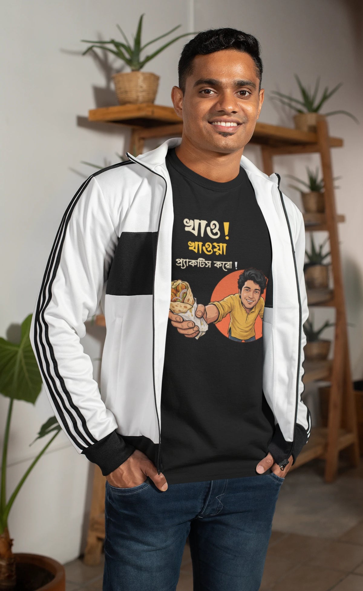 A smiling man wearing a "Bong Cazuals" graphic t-shirt featuring Bengali typography and an illustrated character offering a food roll, shot in an indoor office or lifestyle setting.