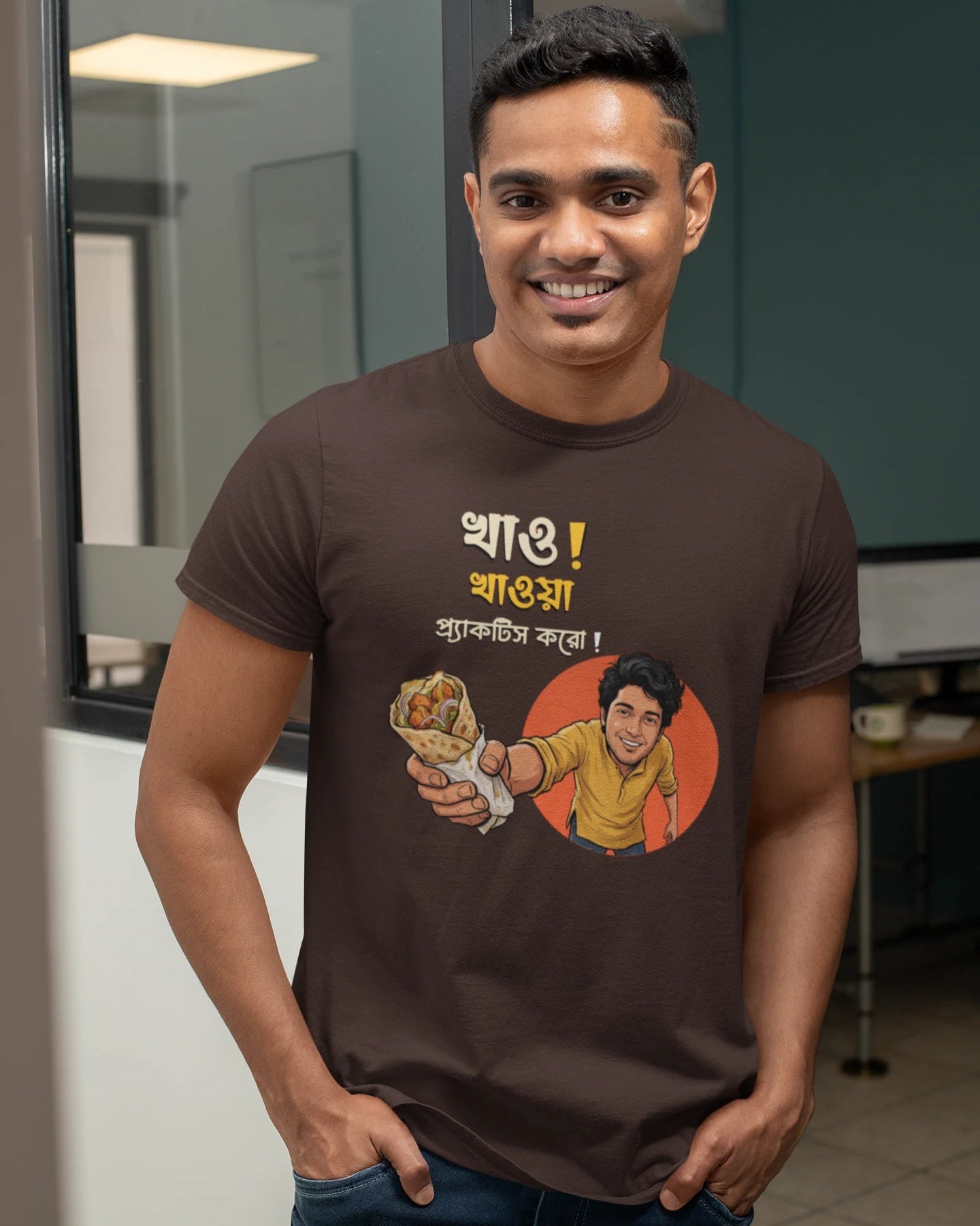 A smiling man wearing a "Bong Cazuals" graphic t-shirt featuring Bengali typography and an illustrated character offering a food roll, shot in an indoor office or lifestyle setting.