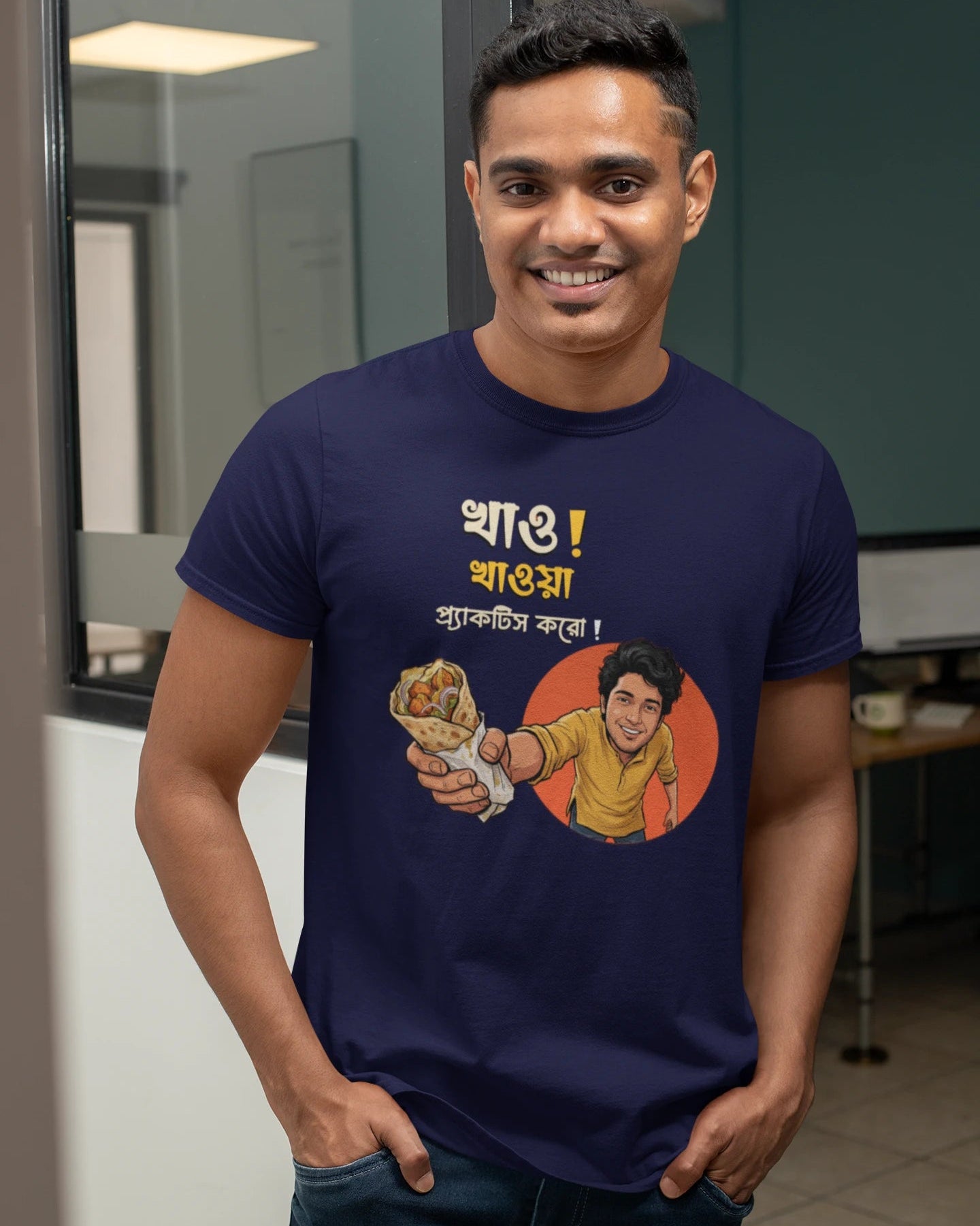 A smiling man wearing a dark "Bong Cazuals" graphic t-shirt featuring Bengali typography and an illustrated character offering a food roll, shot in an indoor office or lifestyle setting.