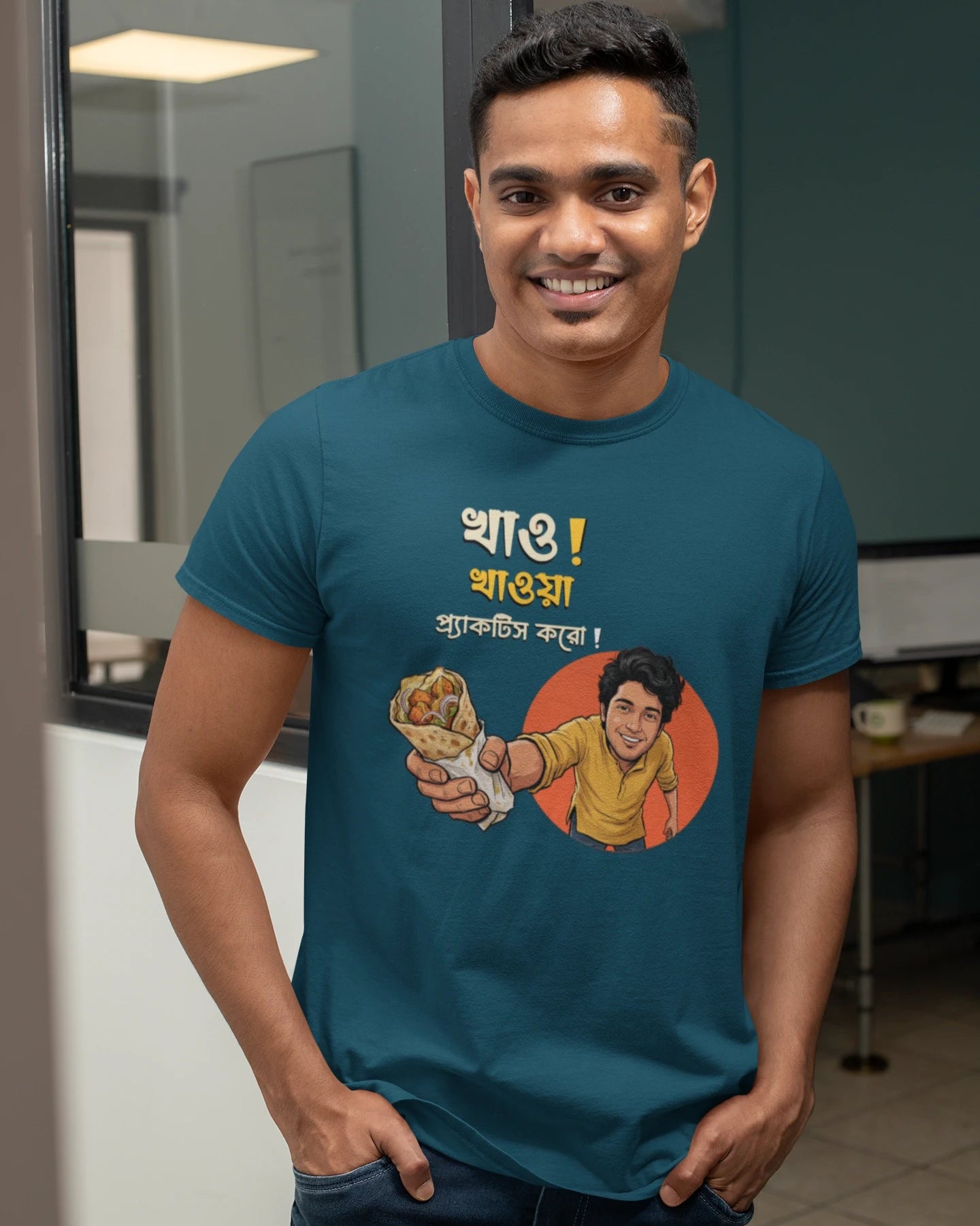 A smiling man wearing a "Bong Cazuals" graphic t-shirt featuring Bengali typography and an illustrated character offering a food roll, shot in an indoor office or lifestyle setting.