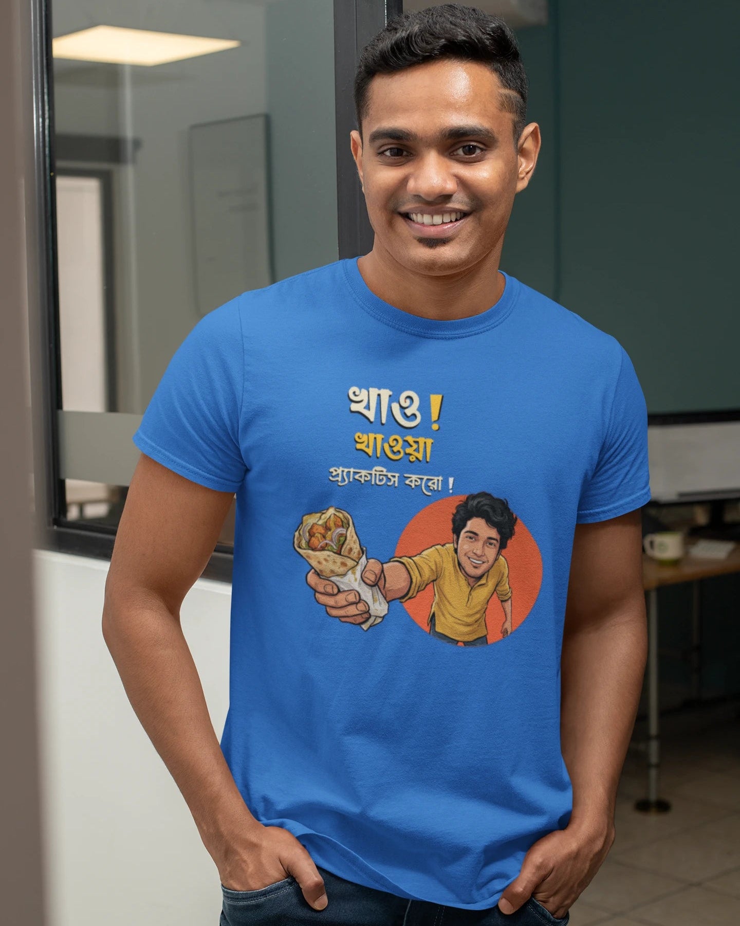 A smiling man wearing a "Bong Cazuals" graphic t-shirt featuring Bengali typography and an illustrated character offering a food roll, shot in an indoor office or lifestyle setting.