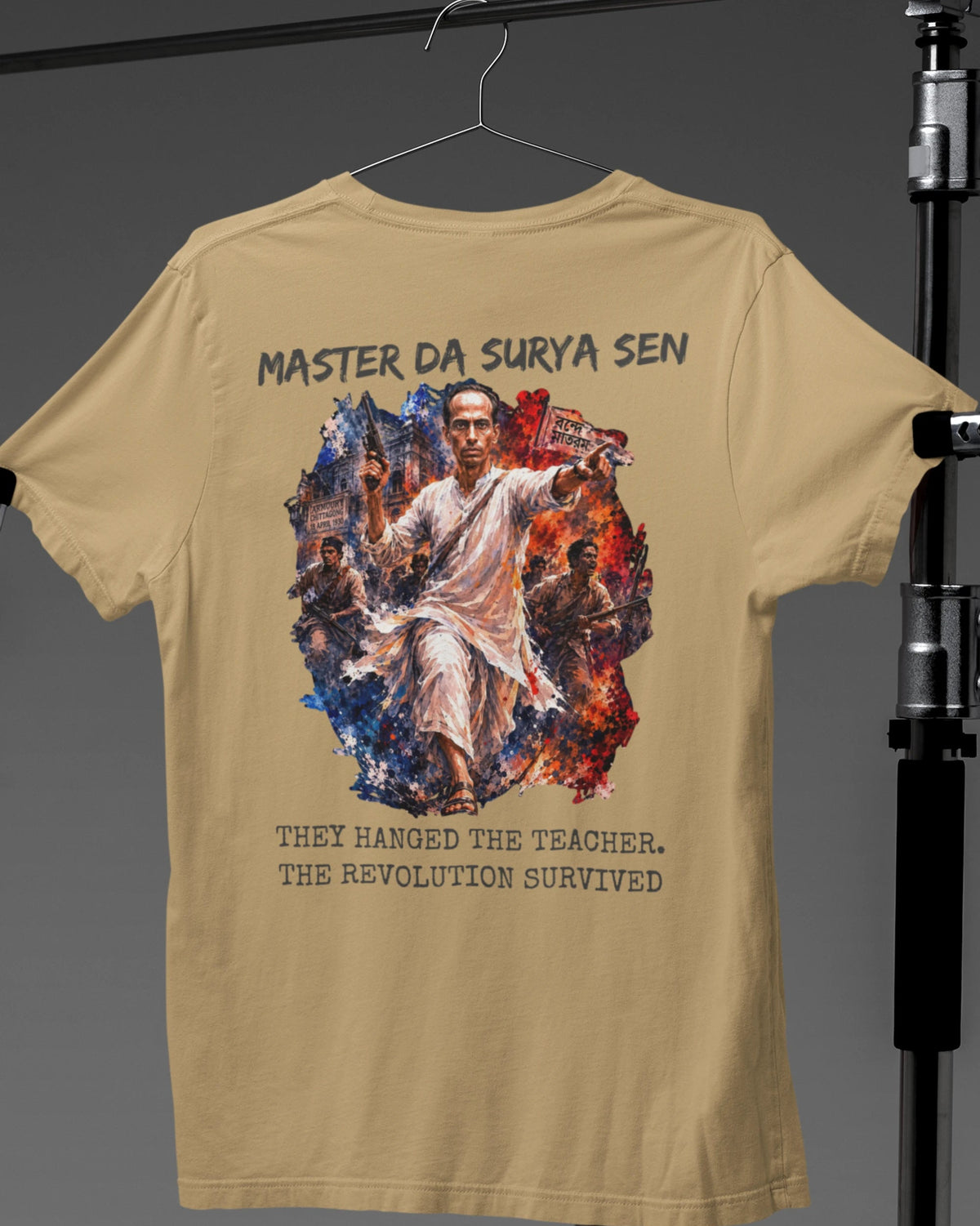A beige graphic t-shirt hanging on a hanger against a dark gray background, featuring a large print of a man surrounded by flames and text that reads 'MASTER DA SURYA SEN' and 'THEY HANGED THE TEACHER, THE REVOLUTION SURVIVED'.