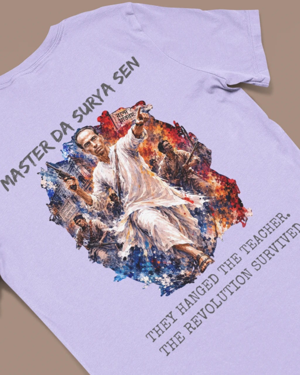 A light lavender-purple t-shirt laid flat on a solid tan background, featuring a large graphic print of a revolutionary figure with the text 'MASTER DA SURYA SEN' and 'THEY HANG THE TEACHER, THE REVOLUTION SURVIVES'.