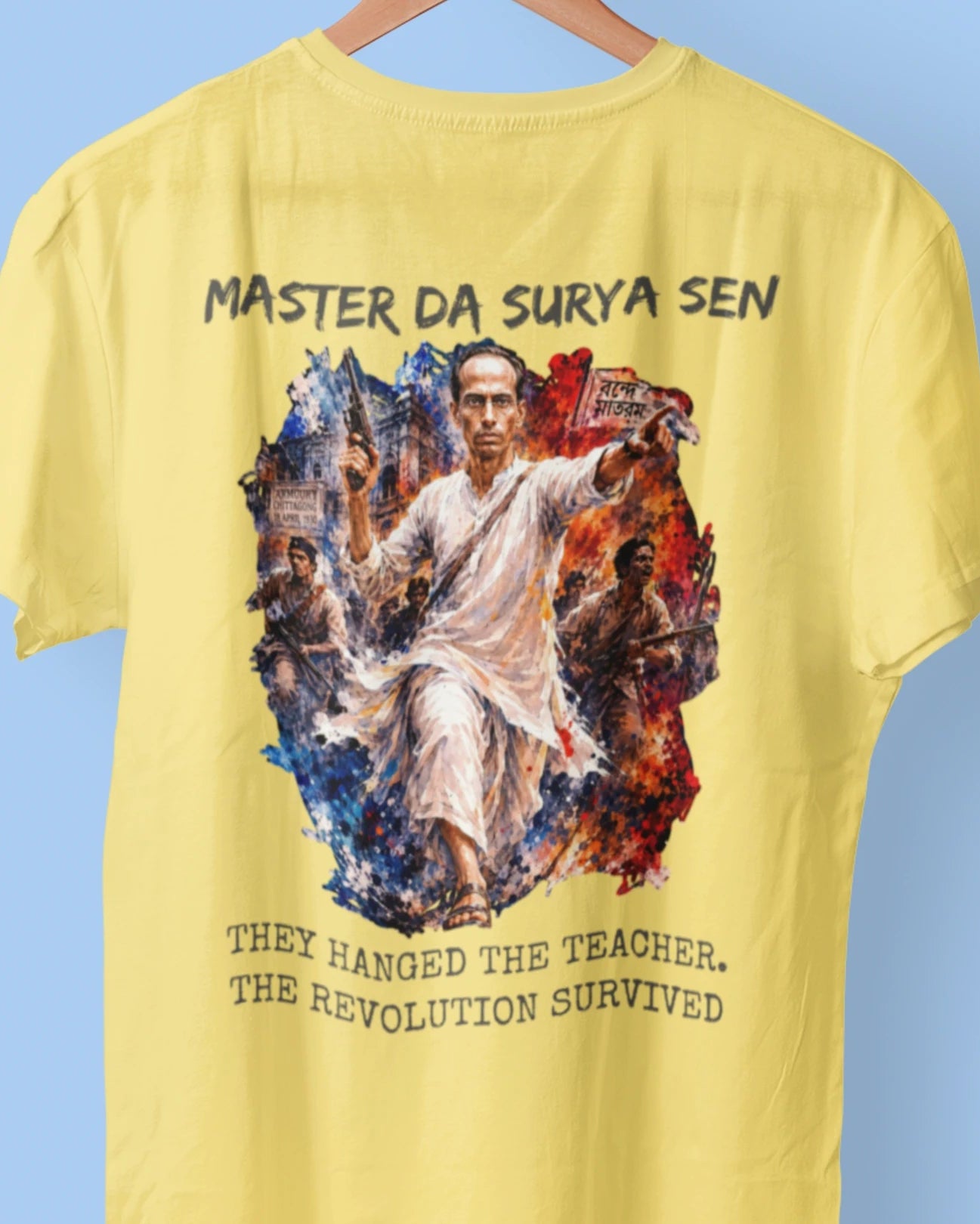 A yellow short-sleeve t-shirt hanging on a wooden hanger against a light blue background. The back of the shirt features a large graphic print of a central figure in a white robe surrounded by a colorful, textured circular border, with the text 'MASTER DA SURYA SEN' above and 'THEY HANGED THE TEACHER.