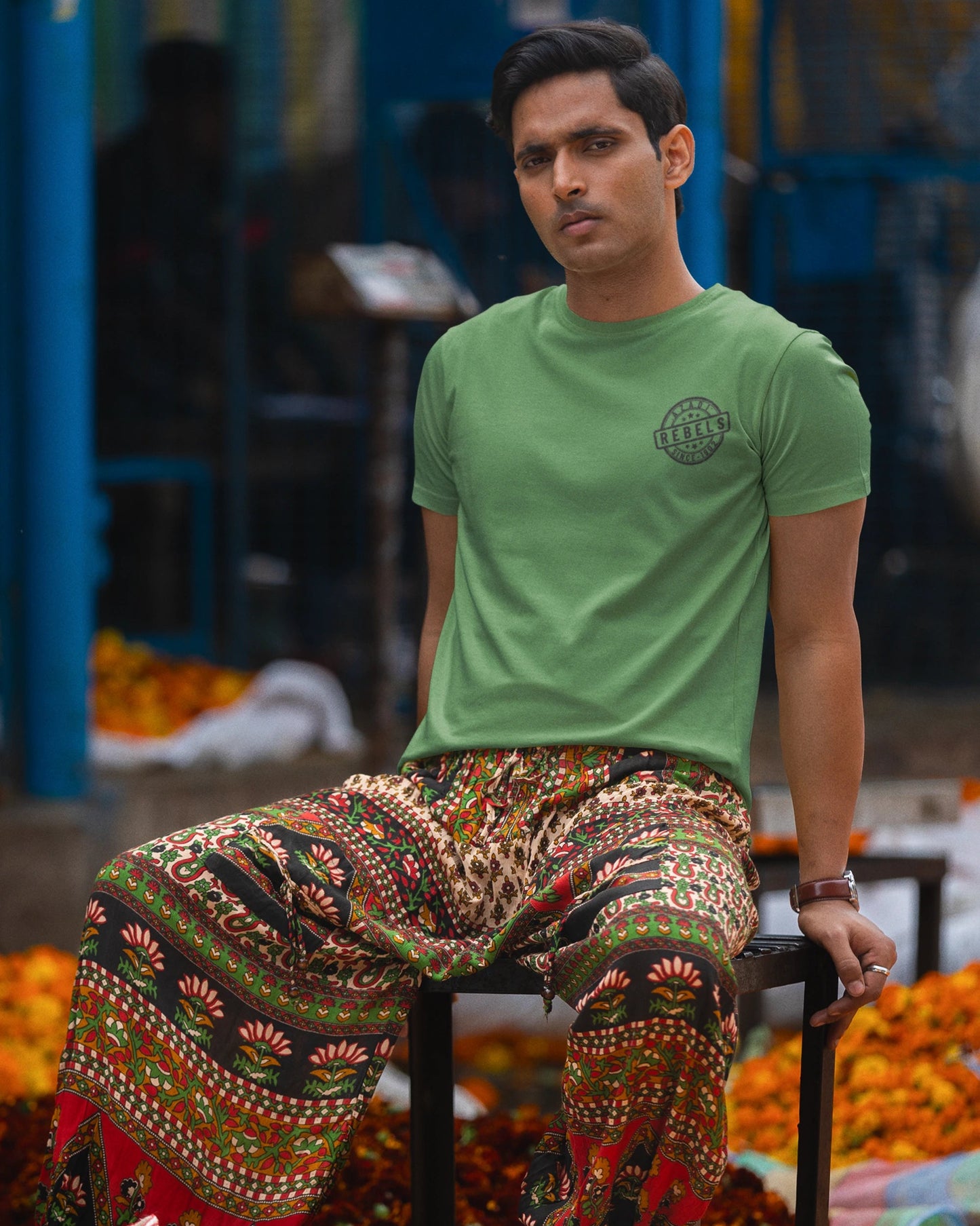 A man sitting on a stool in a market setting, wearing a light green crew-neck t-shirt with a small circular graphic on the left chest and highly patterned, multicolored wide-leg pants.