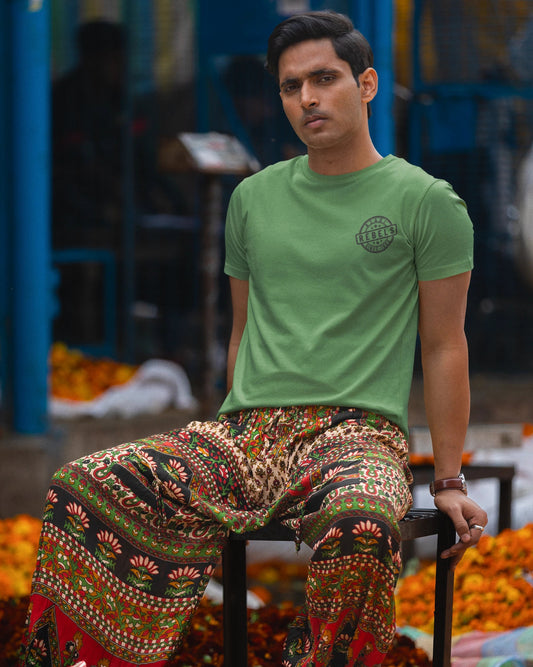 A man sitting on a stool in a market setting, wearing a light green crew-neck t-shirt with a small circular graphic on the left chest and highly patterned, multicolored wide-leg pants.