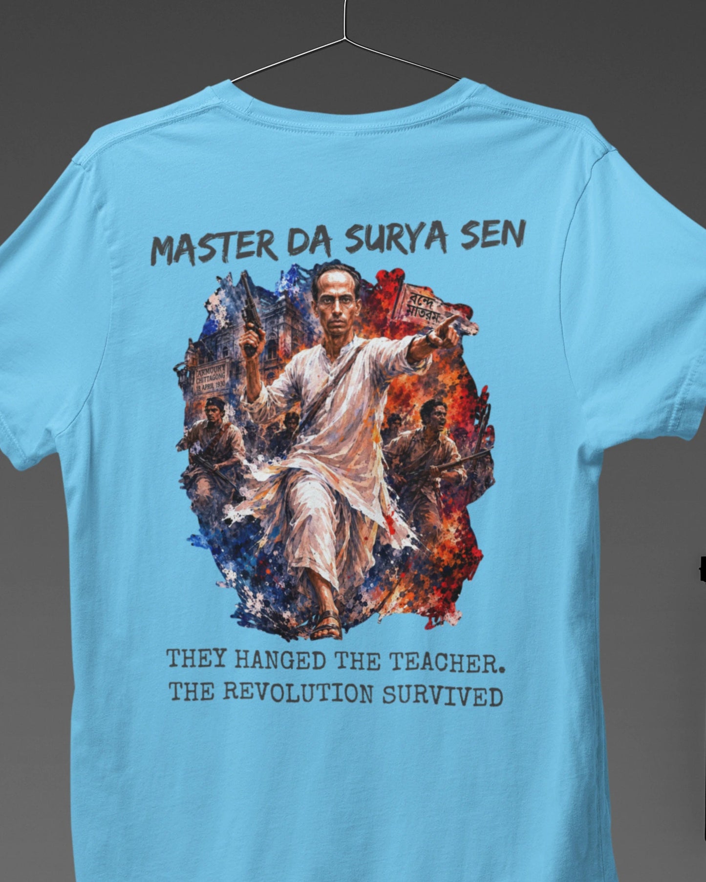 A light blue short-sleeve t-shirt displayed on a hanger against a dark gray background, showing the back with a large graphic print of a man surrounded by flames and text reading 'MASTER DA SURYA SEN' and 'THEY HANGED THE TEACHER. THE REVOLUTION SURVIVED'.