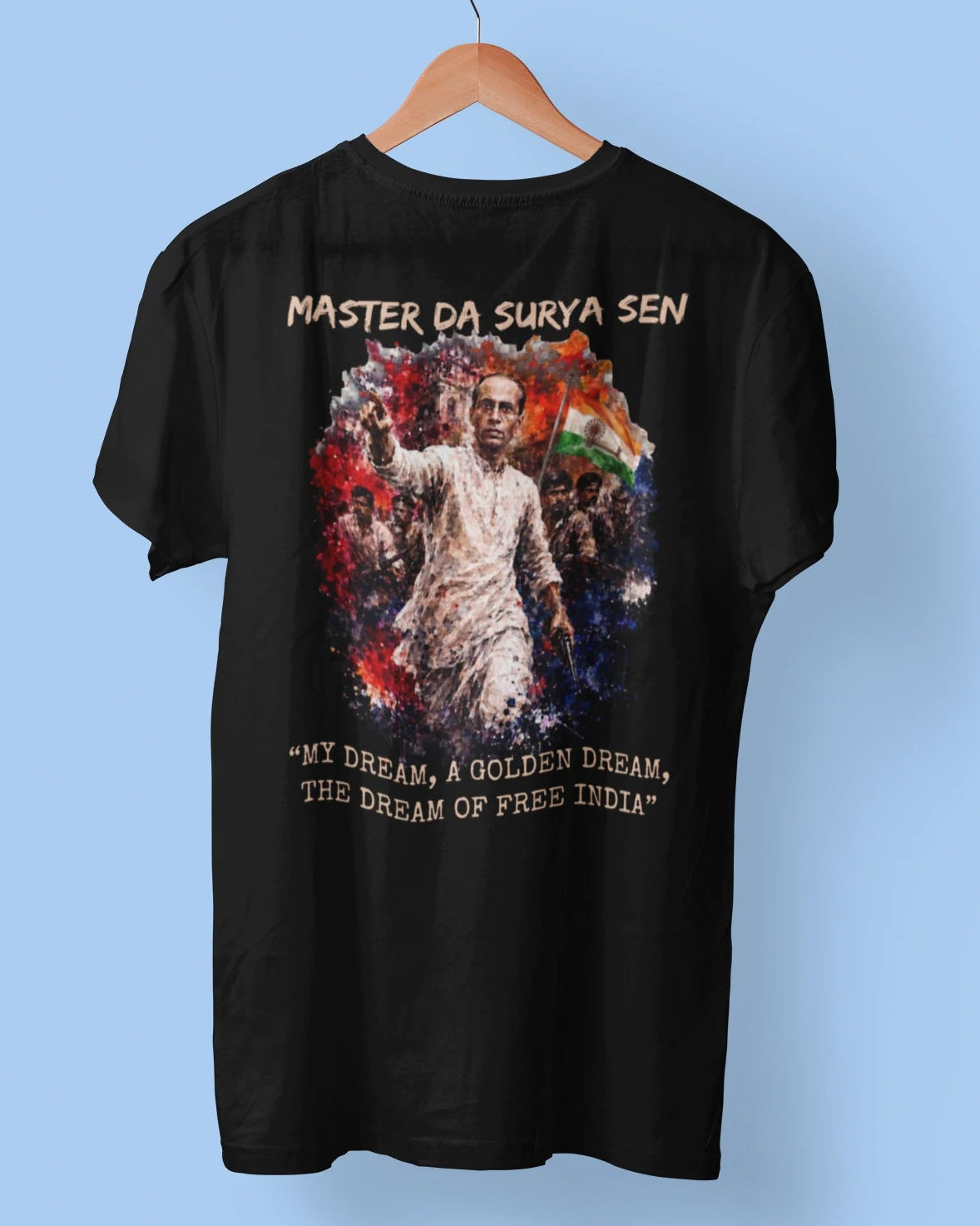 A black short-sleeve graphic t-shirt hanging on a wooden hanger against a light blue background. The shirt features a central graphic of a man in a white kurta surrounded by red, white, and green elements, with the text 'MASTER DA SURYA SEN' above and 'MY DREAM, A GOLDEN DREAM, THE DREAM OF FREE INDIA' below.