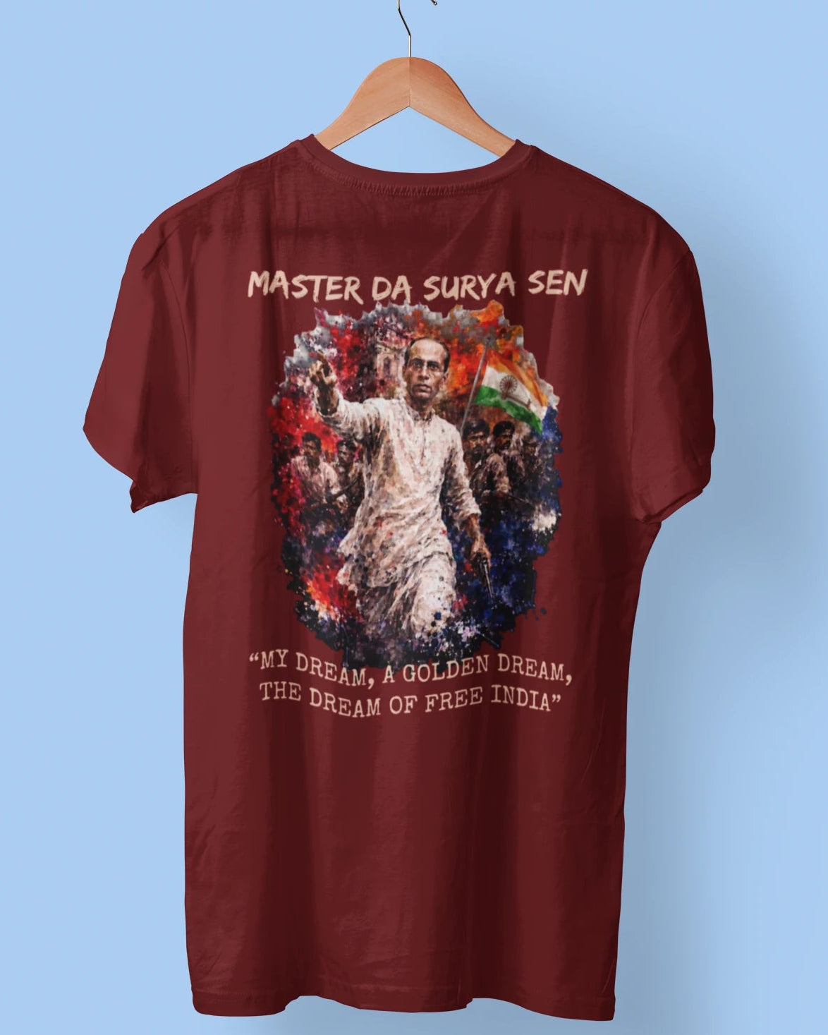 A maroon short-sleeve t-shirt hanging on a wooden hanger against a light blue background. The shirt features a large graphic print on the front depicting a man in a white kurta with an Indian flag, surrounded by red, white, and blue artistic elements, with text reading 'MASTER DA SURYA SEN' and '"MY DREAM A GOLDEN DREAM, THE DREAM OF FREE INDIA"'.