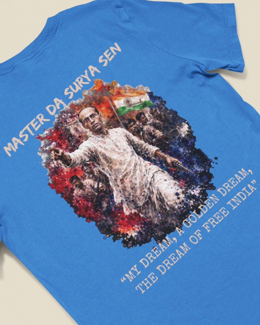 A blue short-sleeve t-shirt laid flat on a light beige background, featuring a large graphic print of a man with the Indian flag, surrounded by text reading 'MASTER DA. SURYA SEN' and 'MY DREAM IS A GOLDEN DREAM, THE DREAM OF FREE INDIA'.