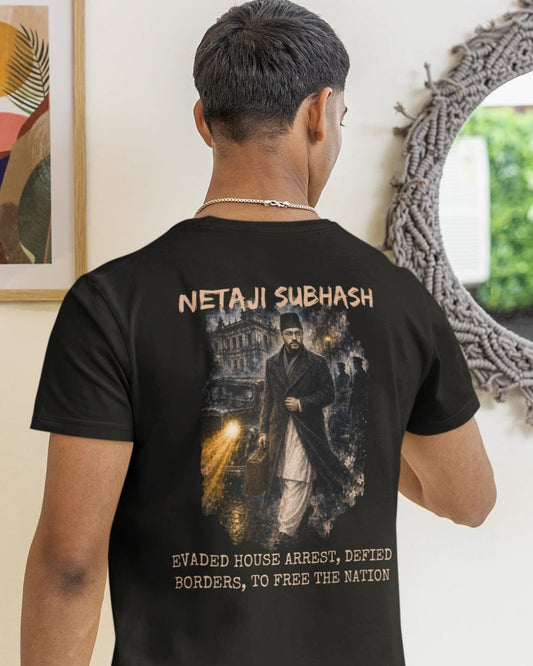 A man viewed from behind wearing a black graphic t-shirt with a large print on the back and light gray sweatpants, standing in a room with a mirror, framed art, and a hanging lamp.
