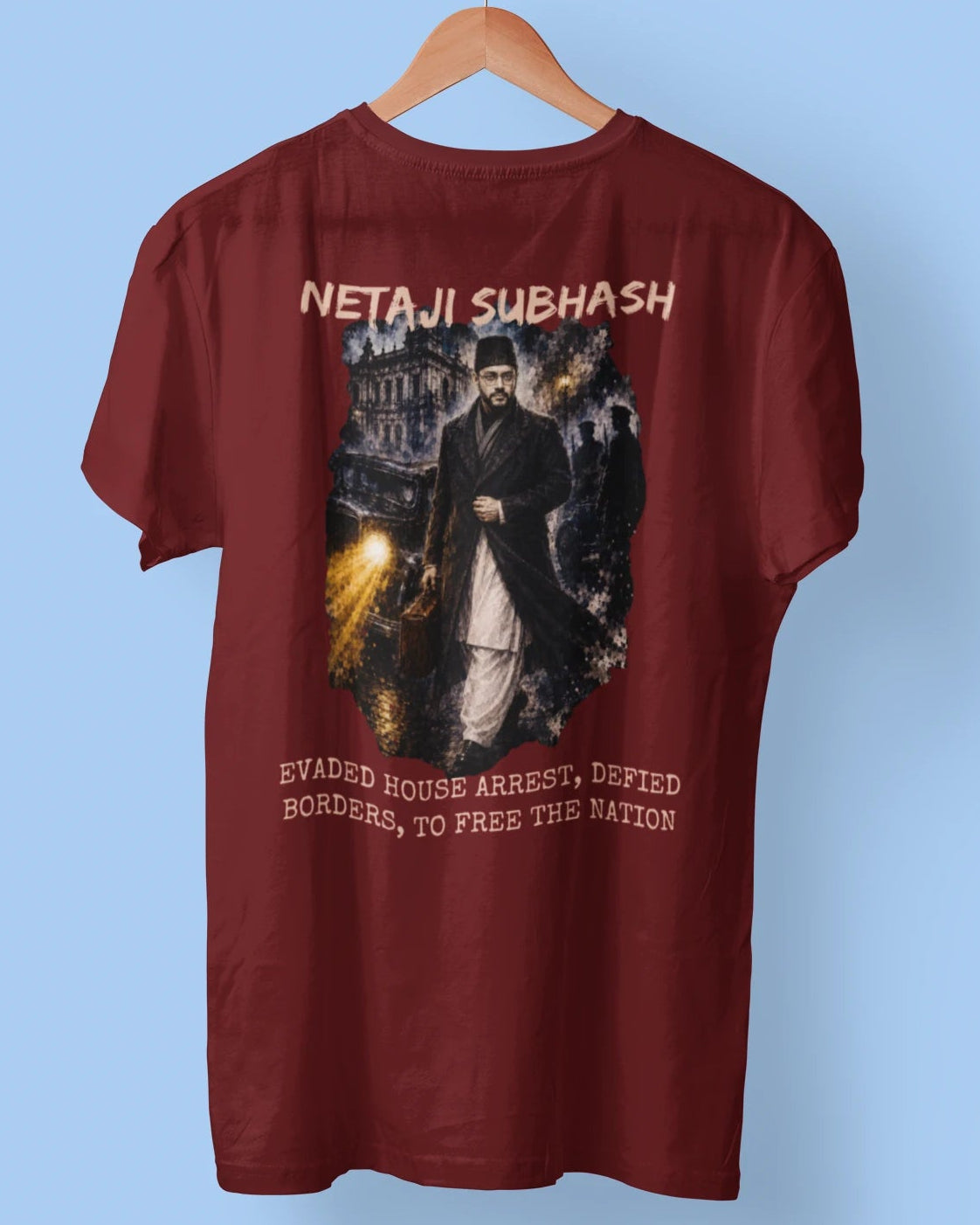 A maroon short-sleeve t-shirt hanging on a wooden hanger against a light blue background. The back of the shirt features a large graphic print with the text 'NETAJI SUBHASH' at the top and 'EWAGED HOUSE ARREST, DEFEATED BORDERS, TO FREE THE NATION' at the bottom, surrounding an illustration of a man in a dark coat.