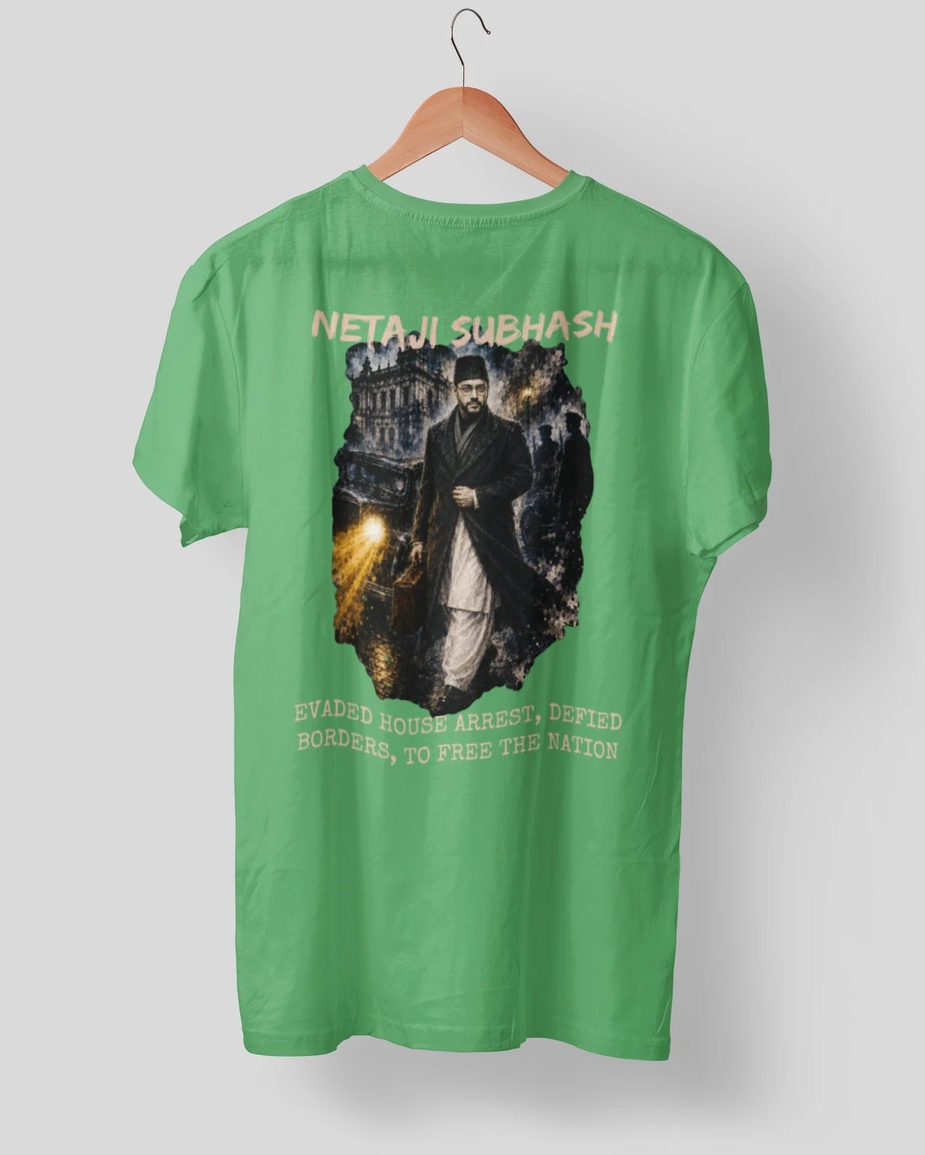A green short-sleeve t-shirt hanging on a wooden hanger against a light gray background. The back of the shirt features a large graphic print depicting a historical figure in a dark suit, with yellow text reading 'NETAJI SUBHASH' above and 'EWAGED HOUSE ARRESTS DEFEATED, BORDERS TO FREE THE NATION' below.