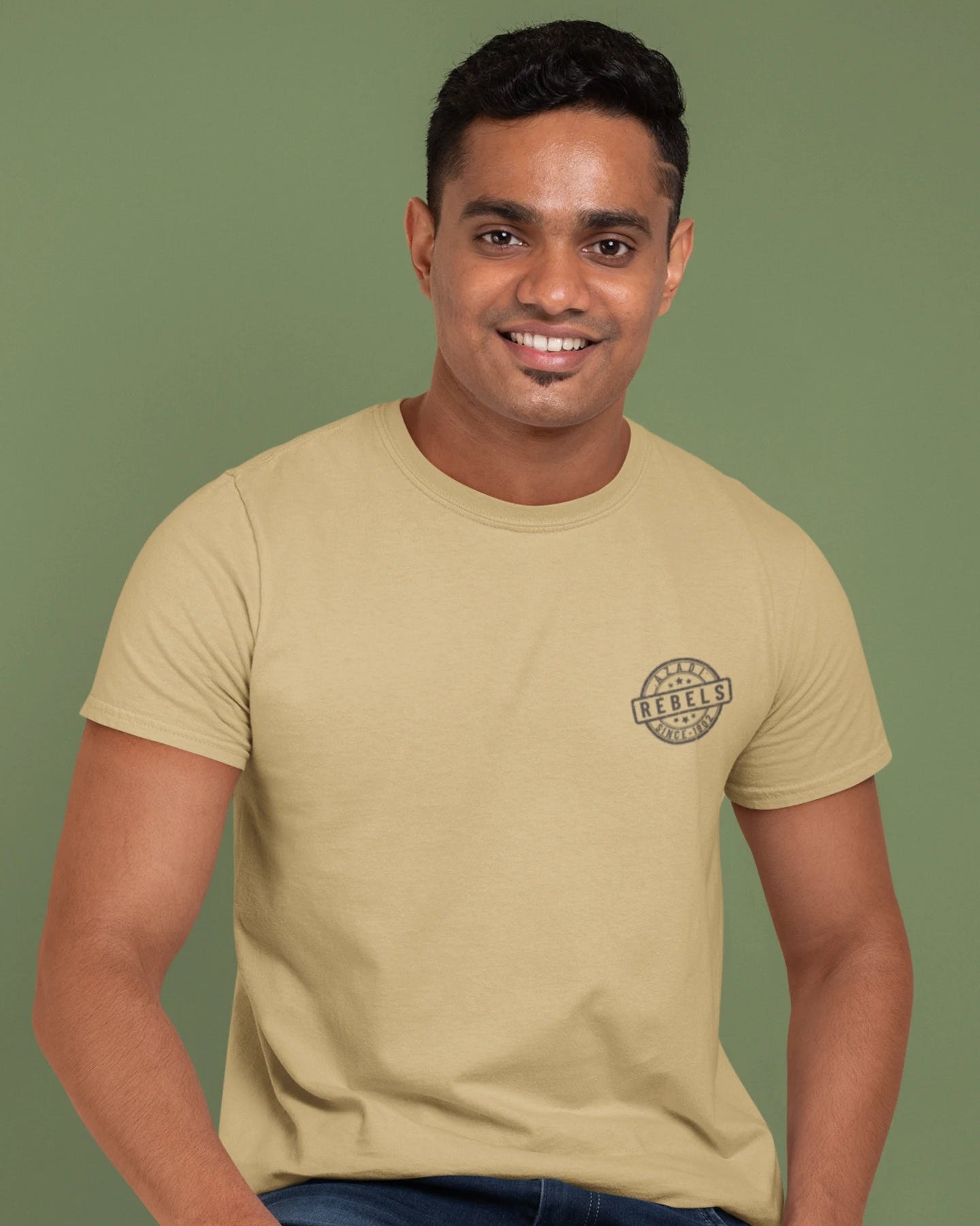 A man wearing a beige crewneck t-shirt with a small circular graphic on the left chest, paired with blue jeans, set against a solid olive green background.