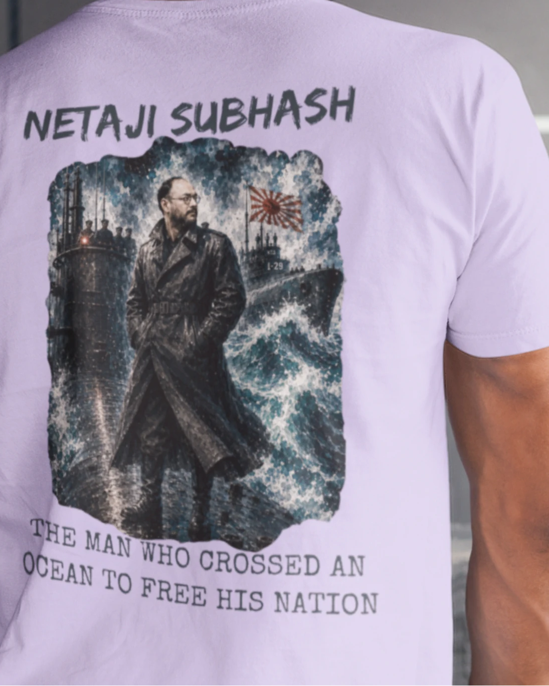 A man viewed from behind wearing a light lavender-purple t-shirt with a large graphic print on the back. The print features the text 'NETAJI SUBHASH' at the top and 'THE MAN WHO CROSSED AN OCEAN TO FREE HIS NATION' at the bottom, surrounding an illustration of a man in a trench coat standing against a dark, industrial background with a Japanese flag.