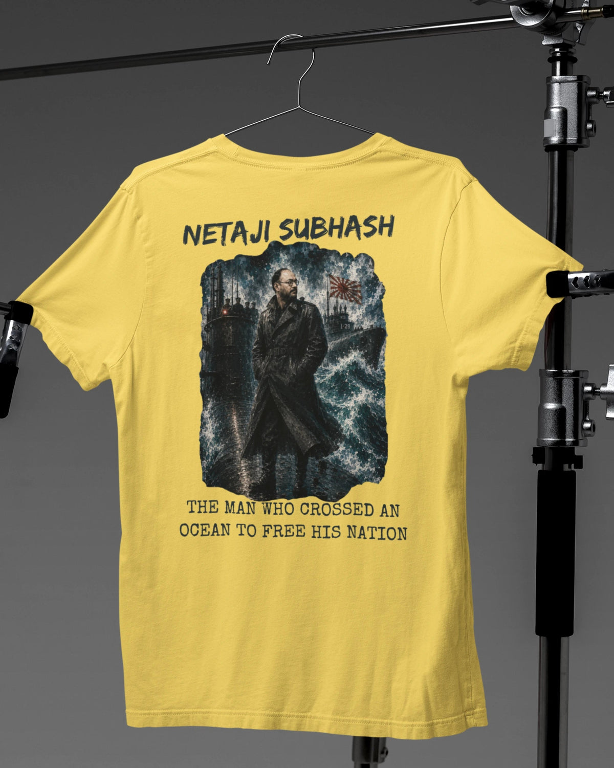 A yellow short-sleeve t-shirt hanging on a hanger against a dark gray studio backdrop, with a large back graphic showing a man in front of stormy waves and the text 'NETAJI SUBHASH' and 'THE MAN WHO CROSSED AN OCEAN TO FREE HIS NATION.' Black clothing stands are visible on both sides.