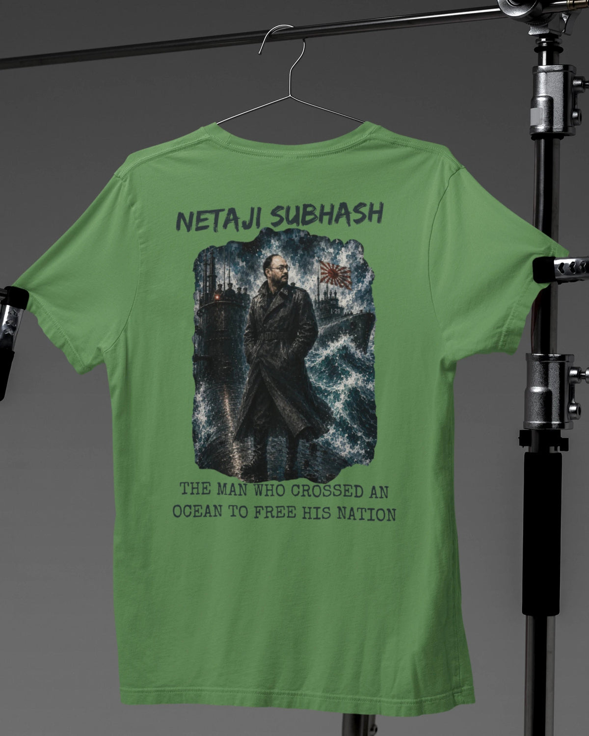 A green short-sleeve T-shirt hanging on a hanger, showing the back side with a large graphic print of Netaji Subhash and text. The shirt is photographed against a dark gray studio background with a light stand visible on both sides.