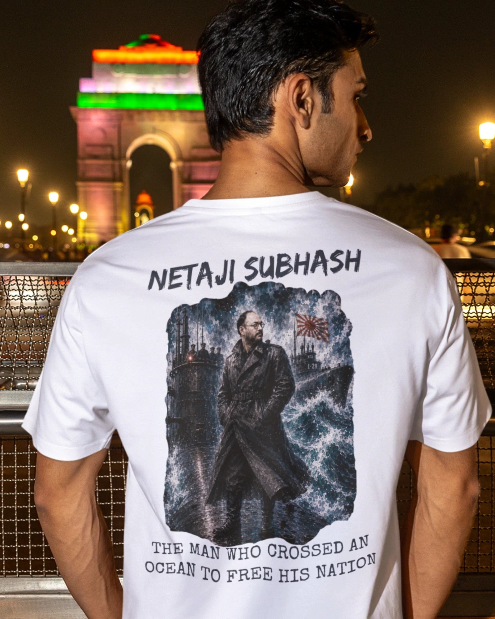 A man wearing a white t-shirt with a large graphic print of Netaji Subhash on the back, standing in front of the India Gate monument at night.