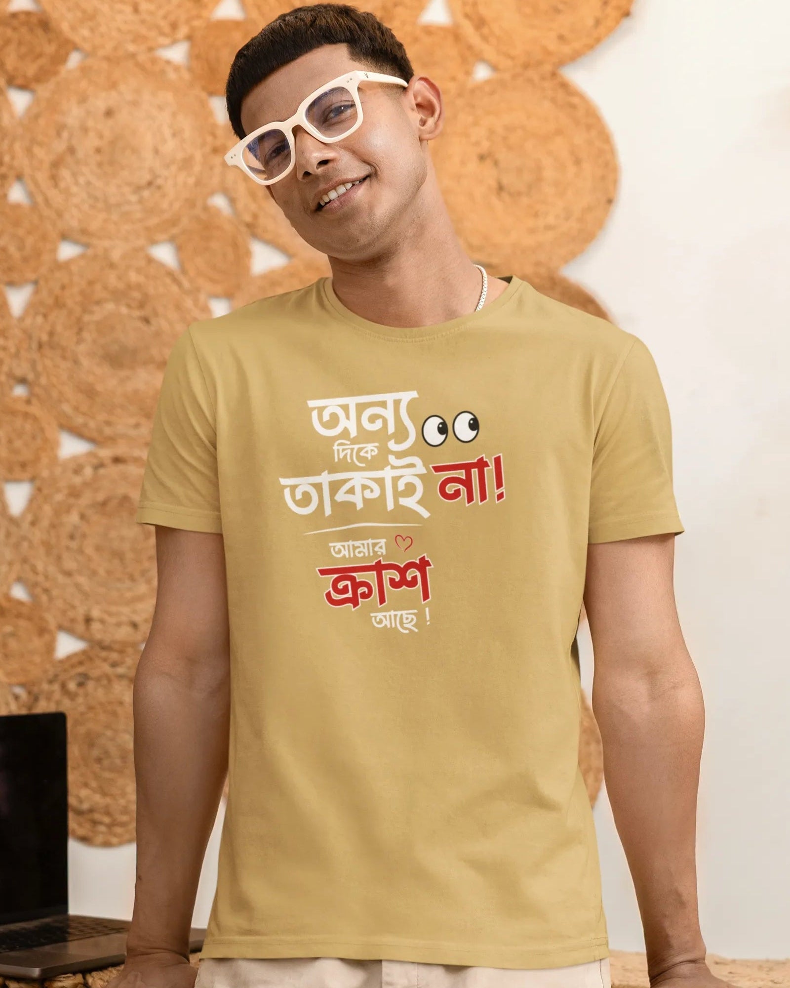 A man wearing a beige graphic t-shirt with Hindi text, white sunglasses, and beige pants, standing in front of a decorative cork wall.