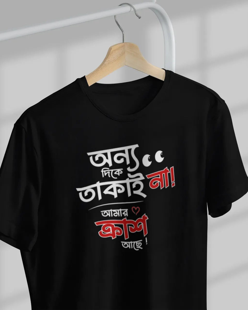A black short-sleeve t-shirt hanging on a wooden hanger against a light gray wall with window shadow patterns. The shirt features a bold graphic print in white and red Bengali text on the front.
