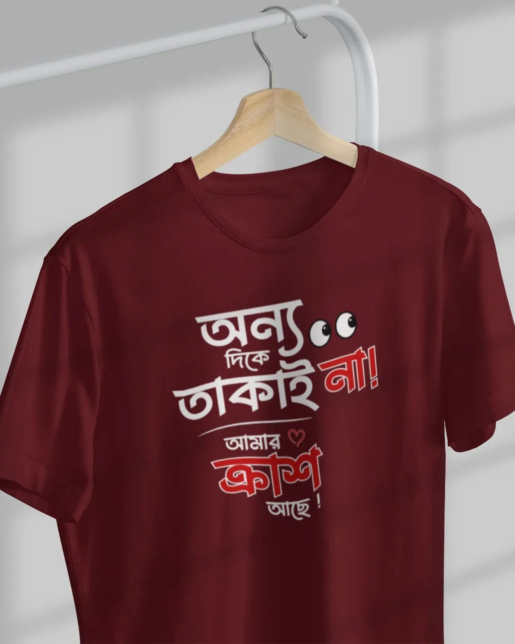 A maroon short-sleeve t-shirt hanging on a wooden hanger against a light gray wall with window shadow patterns. The shirt features Bengali text printed in white and red on the front.