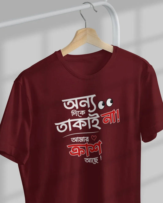A maroon short-sleeve t-shirt hanging on a wooden hanger against a light gray wall with window shadow patterns. The shirt features Bengali text printed in white and red on the front.