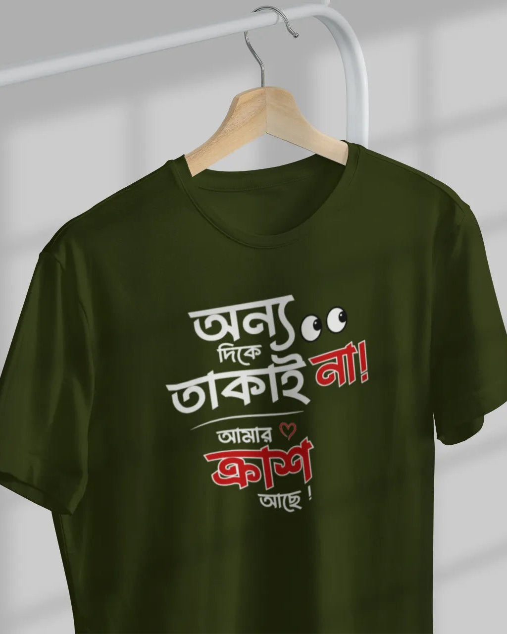 An olive green short-sleeve t-shirt hanging on a wooden hanger against a gray wall with window shadow patterns. The shirt features a bold Bengali text graphic in white and red on the front.