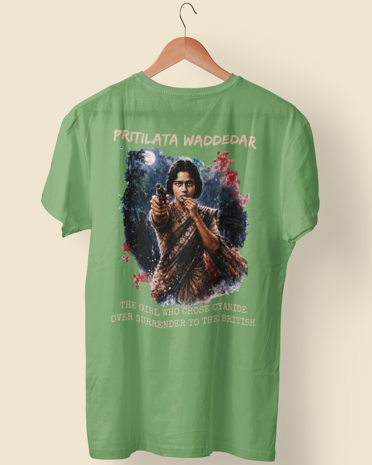A green short-sleeve t-shirt hanging on a wooden hanger against a light beige background. The back of the shirt features a large graphic print of a man holding a gun, surrounded by text that reads 'PRITILATA WADDEDAR' at the top and 'THE GREAT BRITISH MAJESTY OVER SCOTLAND TO THE BRITISH!' at the bottom.