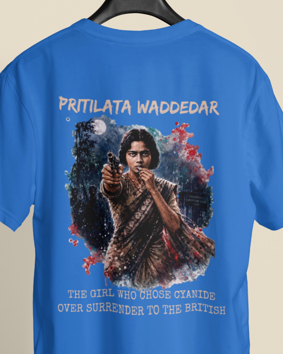A blue short-sleeve t-shirt hanging on a black hanger against a light beige background. The back of the shirt features a large graphic print of a young girl aiming a gun, surrounded by a dark wolf and red splatter details, with the text 'PRITILATA WADDEDAR' above and 'THE GIRL WHO CHOSE CYANIDE OVER SURRENDER TO THE BRITISH' below.