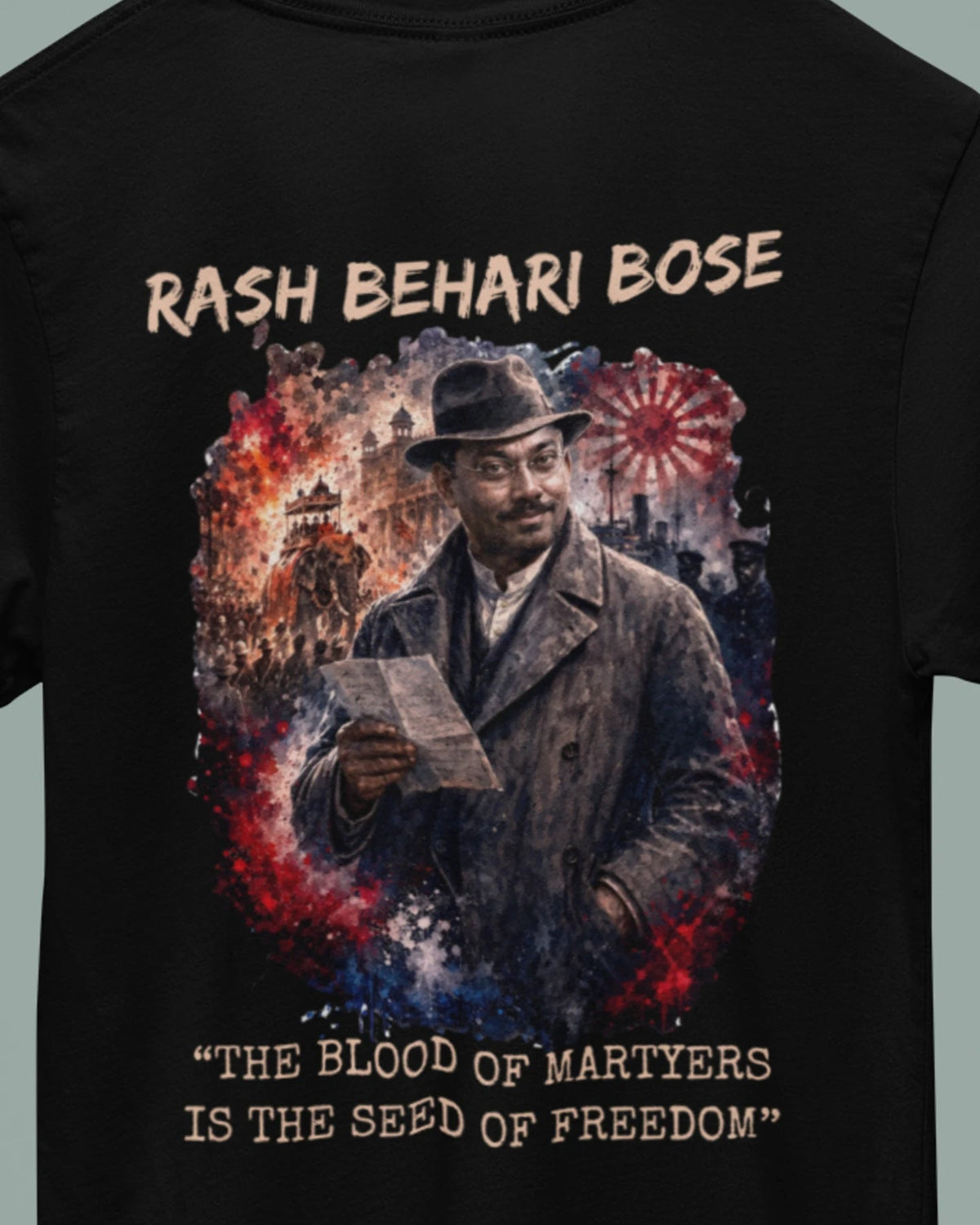 A black short-sleeve t-shirt hanging on a wooden hanger against a muted gray-green background. The back of the shirt features a large graphic print of a man in a hat and coat holding a document, surrounded by red, white, and blue painterly effects, with the text 'RASH BEHARI BOSE' above and 'THE BLOOD OF MARTYERS IS THE SEED OF FREEDOM' below.