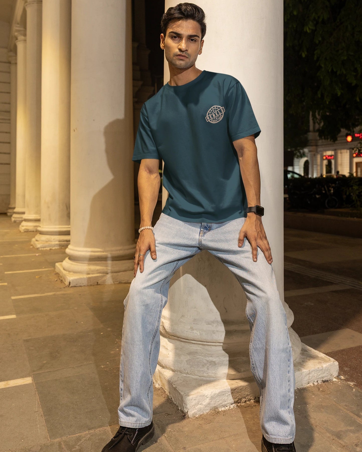 A man posing in a teal blue t-shirt with a circular graphic on the left chest, paired with light wash jeans and black shoes, standing in front of a large white column at night.